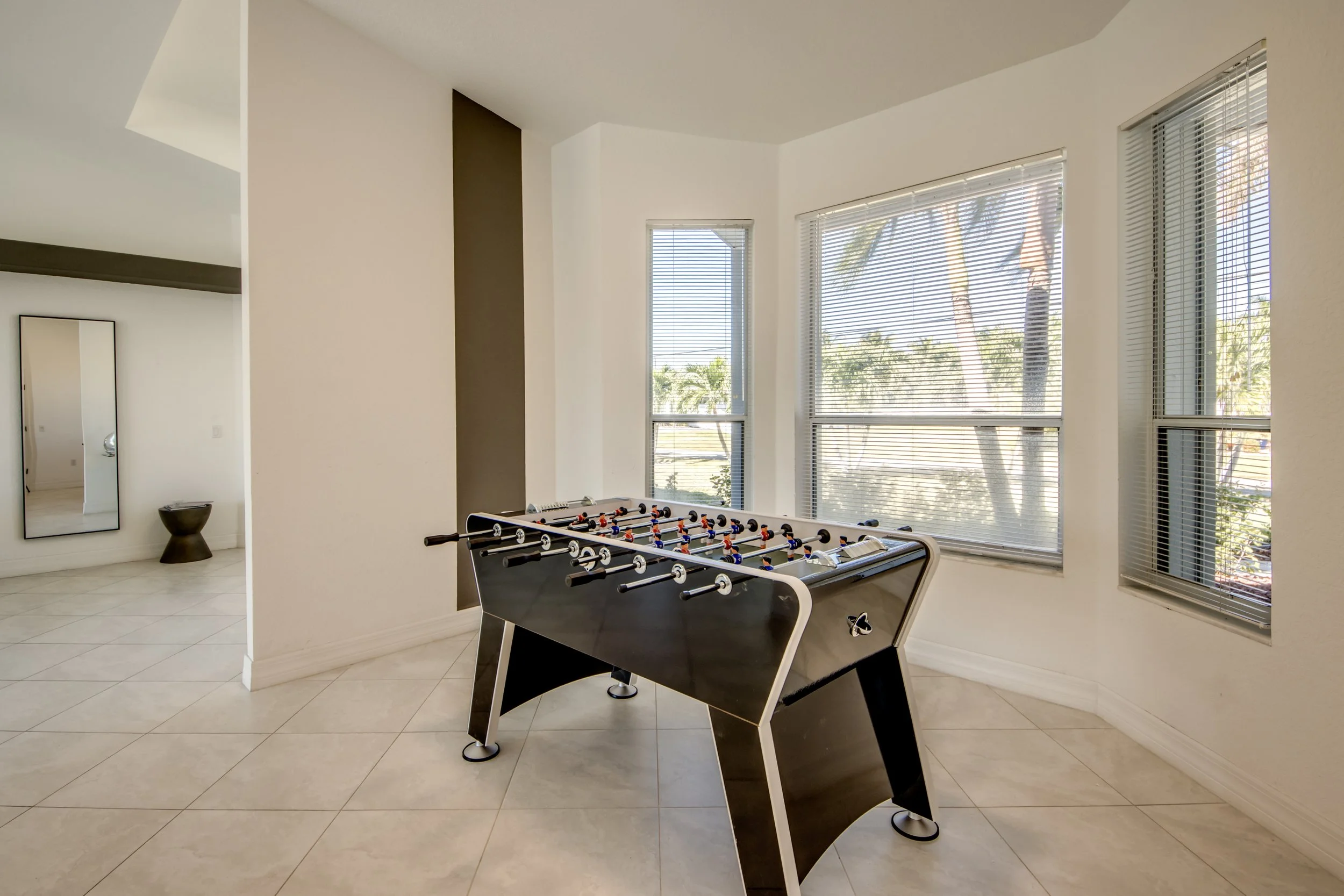 A modern living room with large windows, a foosball table, and minimal decor including a mirror and a small stool.