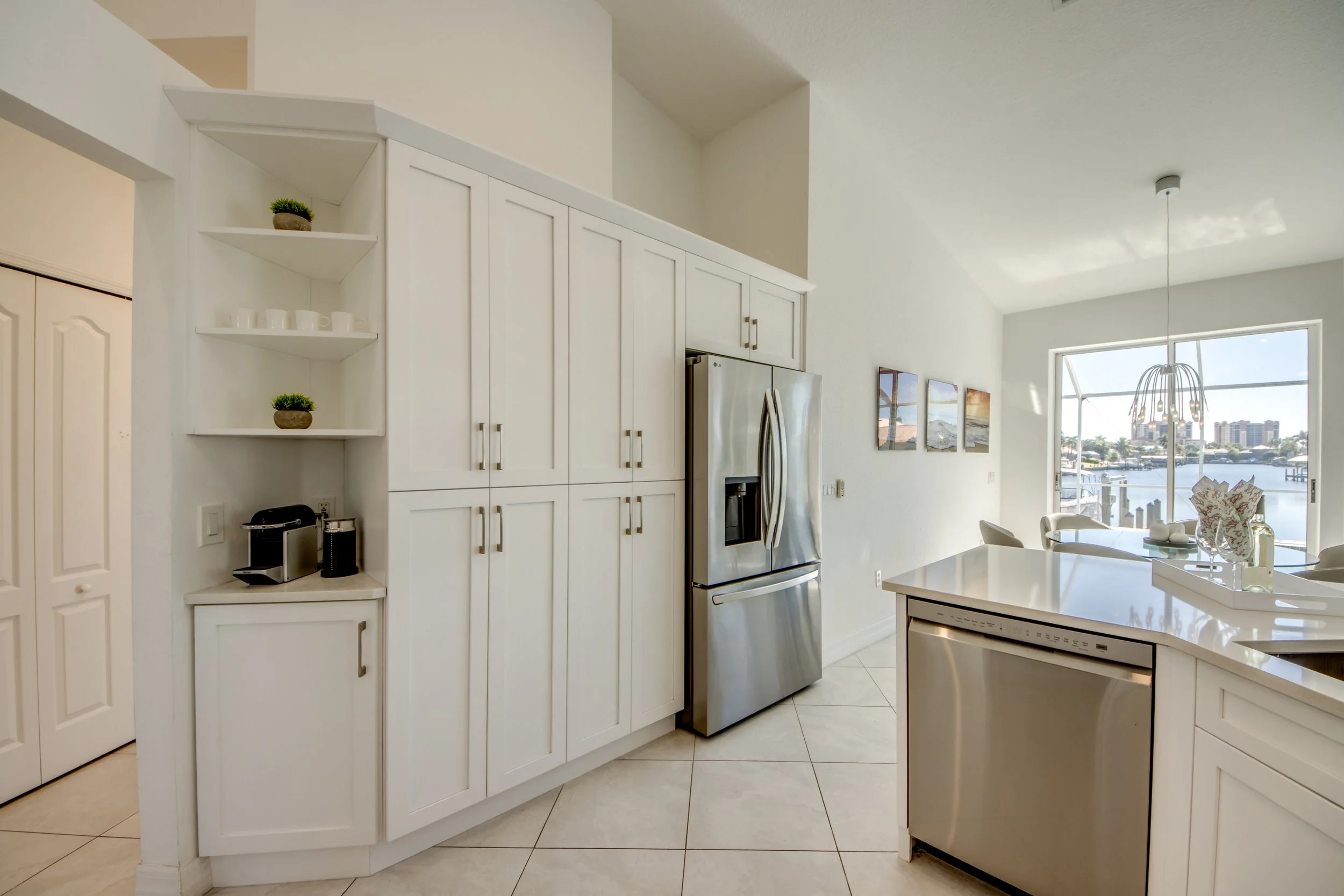 Bright kitchen with white cabinets, stainless steel refrigerator, and large window overlooking water and skyline, with white island and minimal decor.