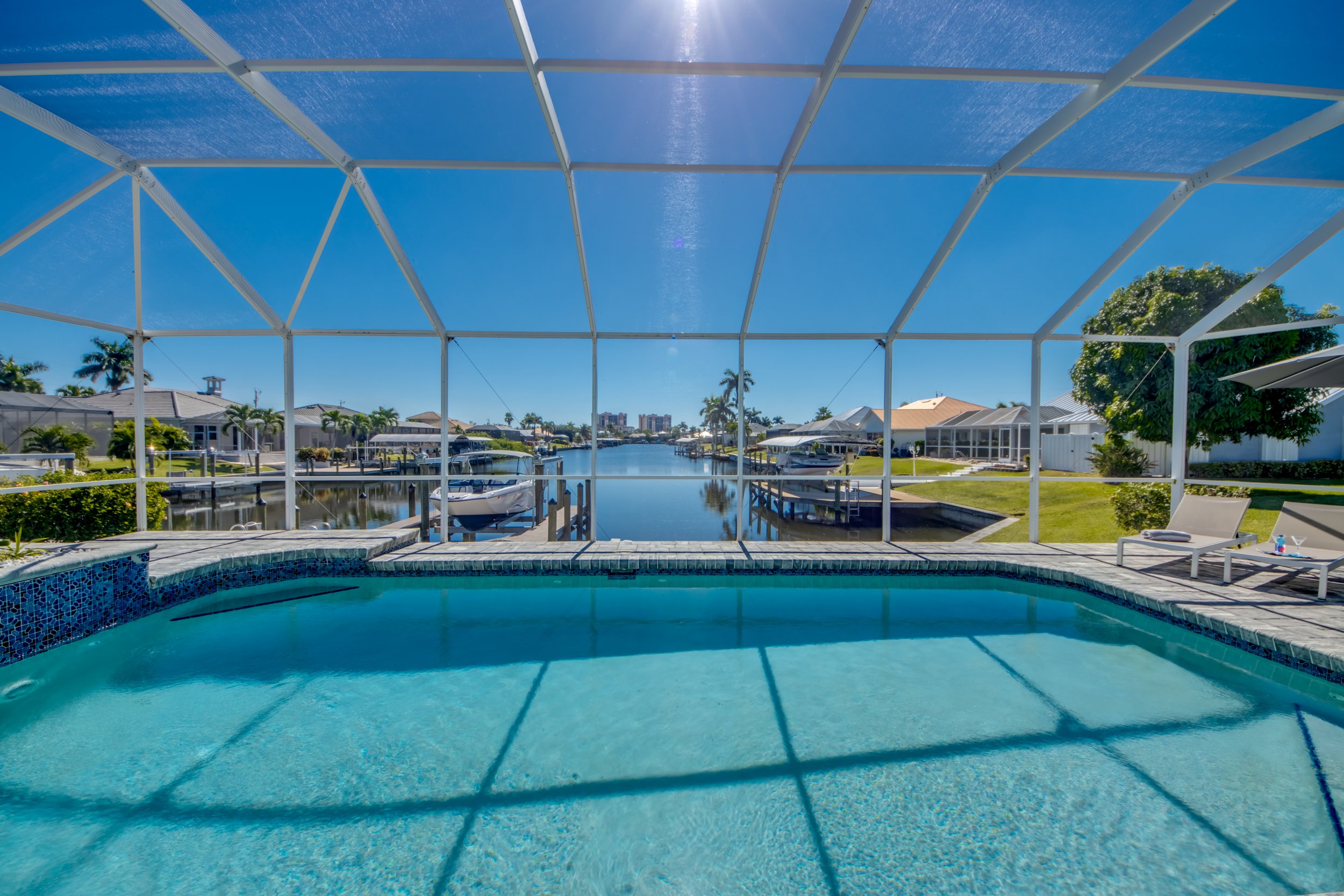 View of a backyard pool enclosed by a screened structure, overlooking a canal with boats, residential houses, and palm trees under a clear blue sky.
