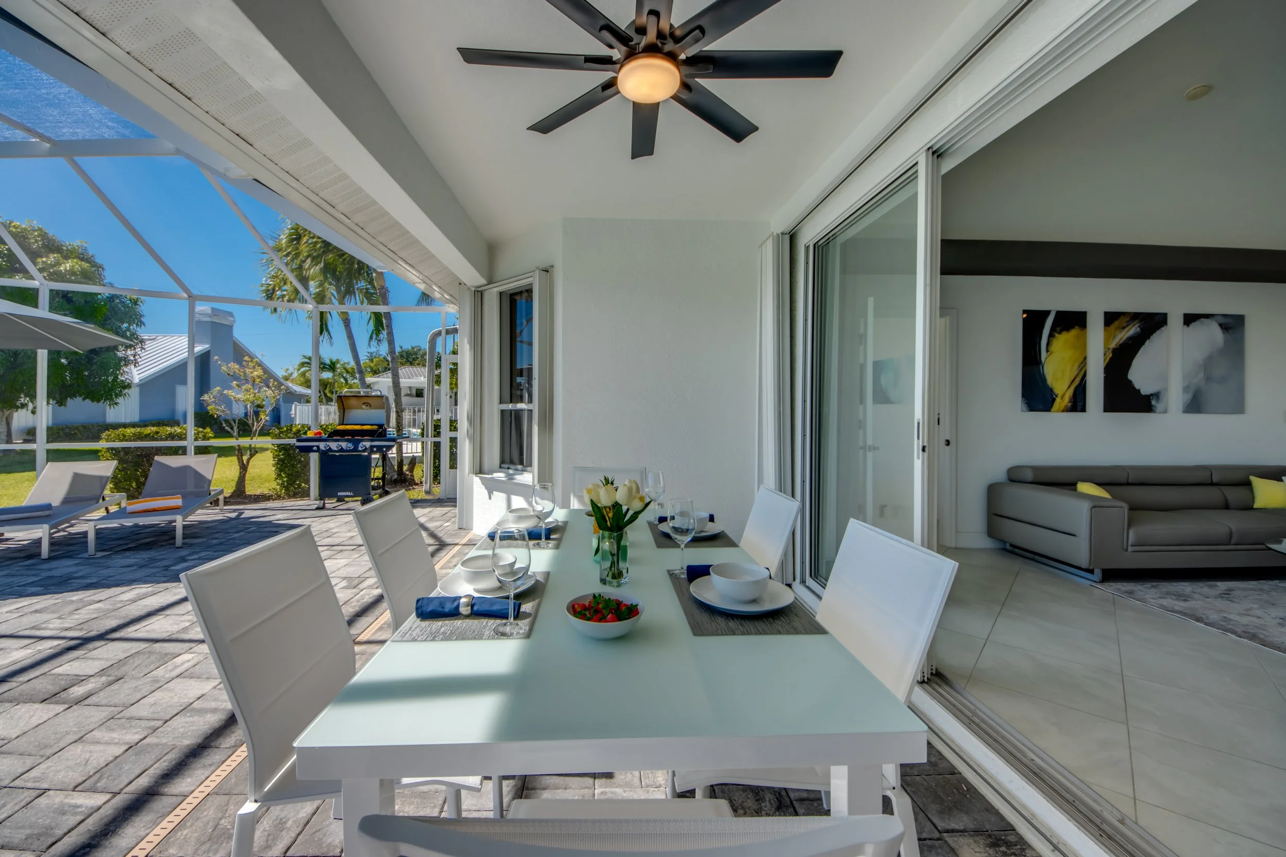 A dining area on a patio with a white table set with dishes, glasses, and a flower arrangement, adjacent to a living room with modern art on the wall, and outdoor furniture including a barbecue grill and lounge chairs visible in the background under 