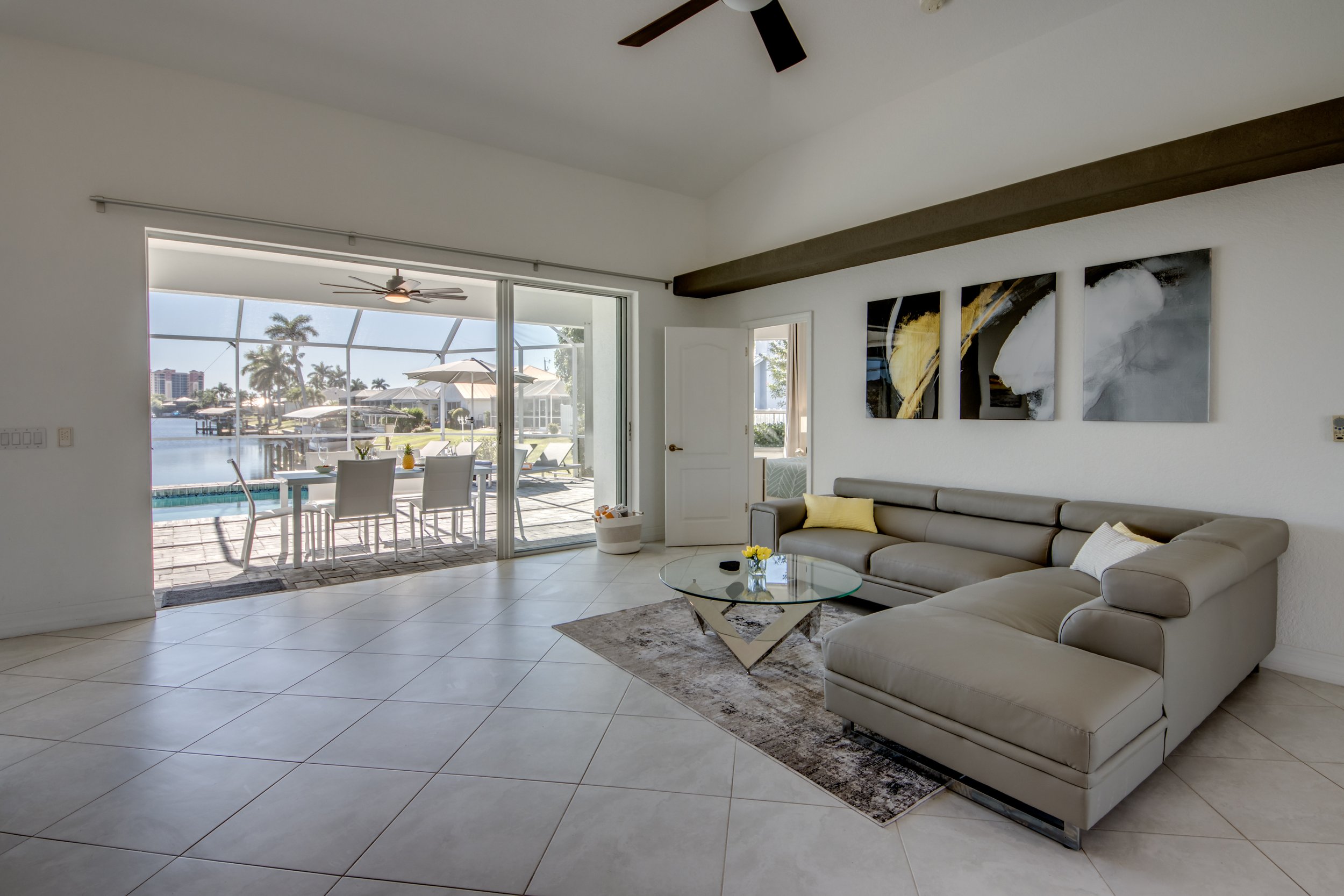 Living room with cream sofa, glass coffee table, and wall art, opening to patio with outdoor dining area, pool, and waterway with boats and palm trees.