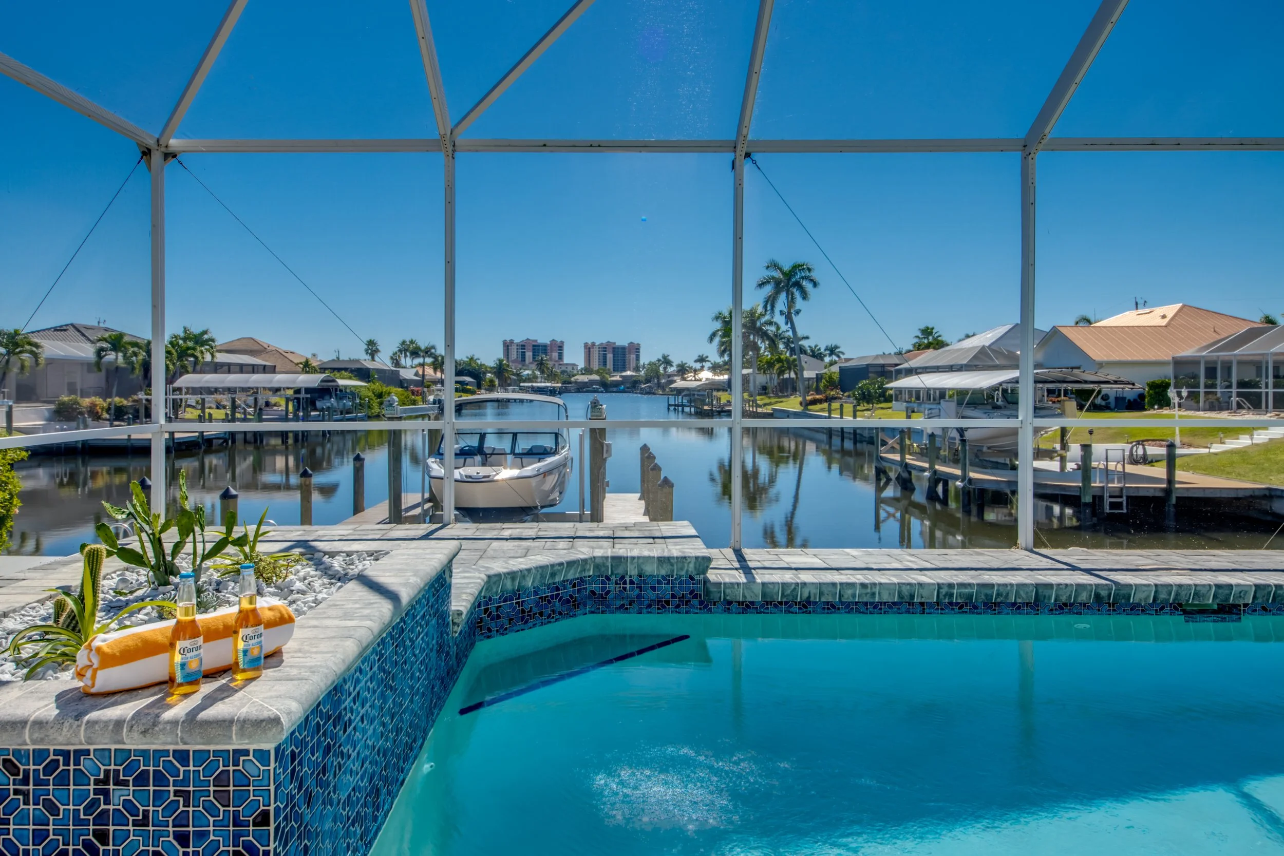 A backyard view of a swimming pool with a hot tub, bottles of Corona beer, and towels on the poolside surrounded by houses, palm trees, and a canal with boats under a clear blue sky.