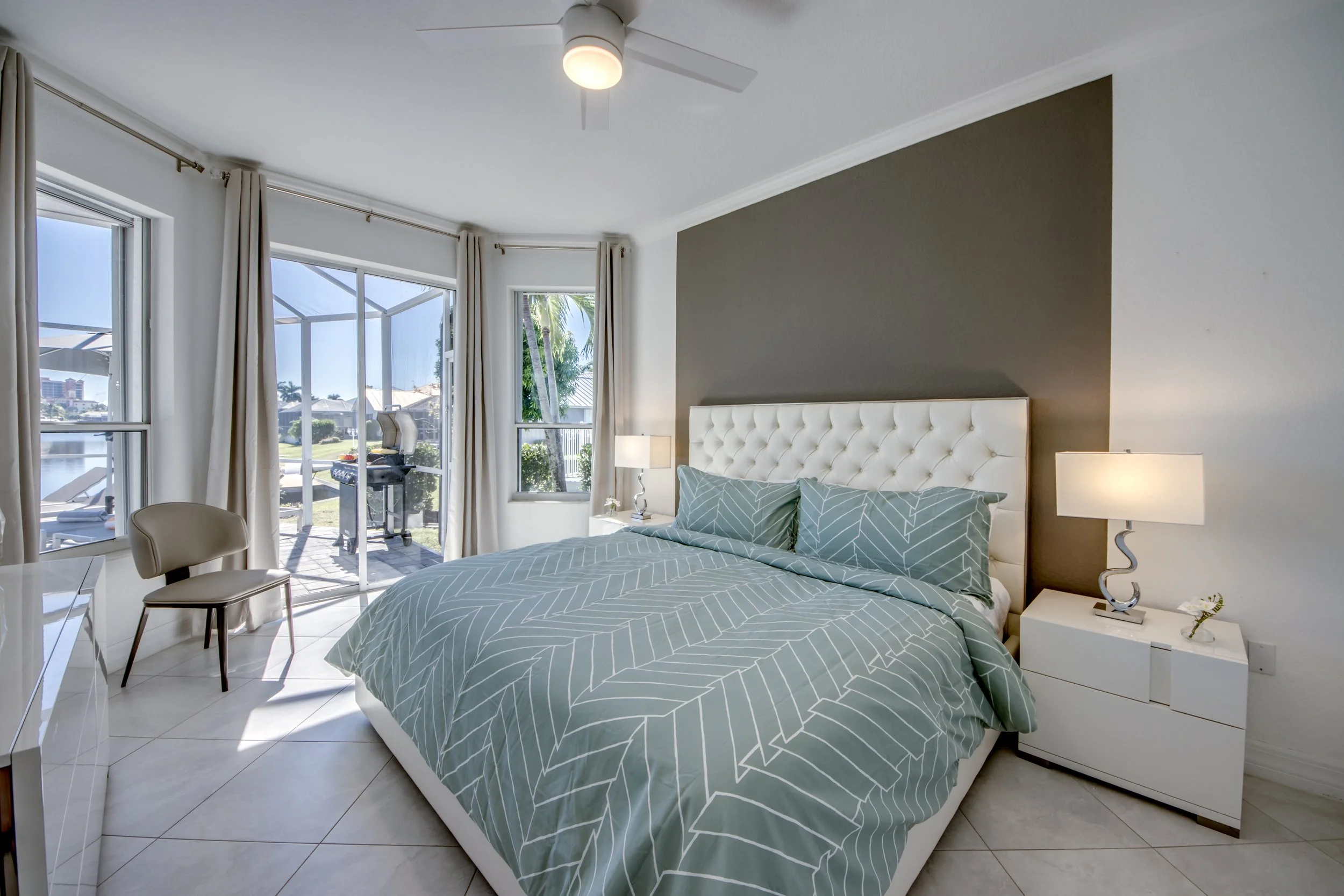 Modern bedroom with large windows, white bed with a gray geometric bedspread, two bedside lamps, and a view of a balcony with outdoor furniture and greenery.
