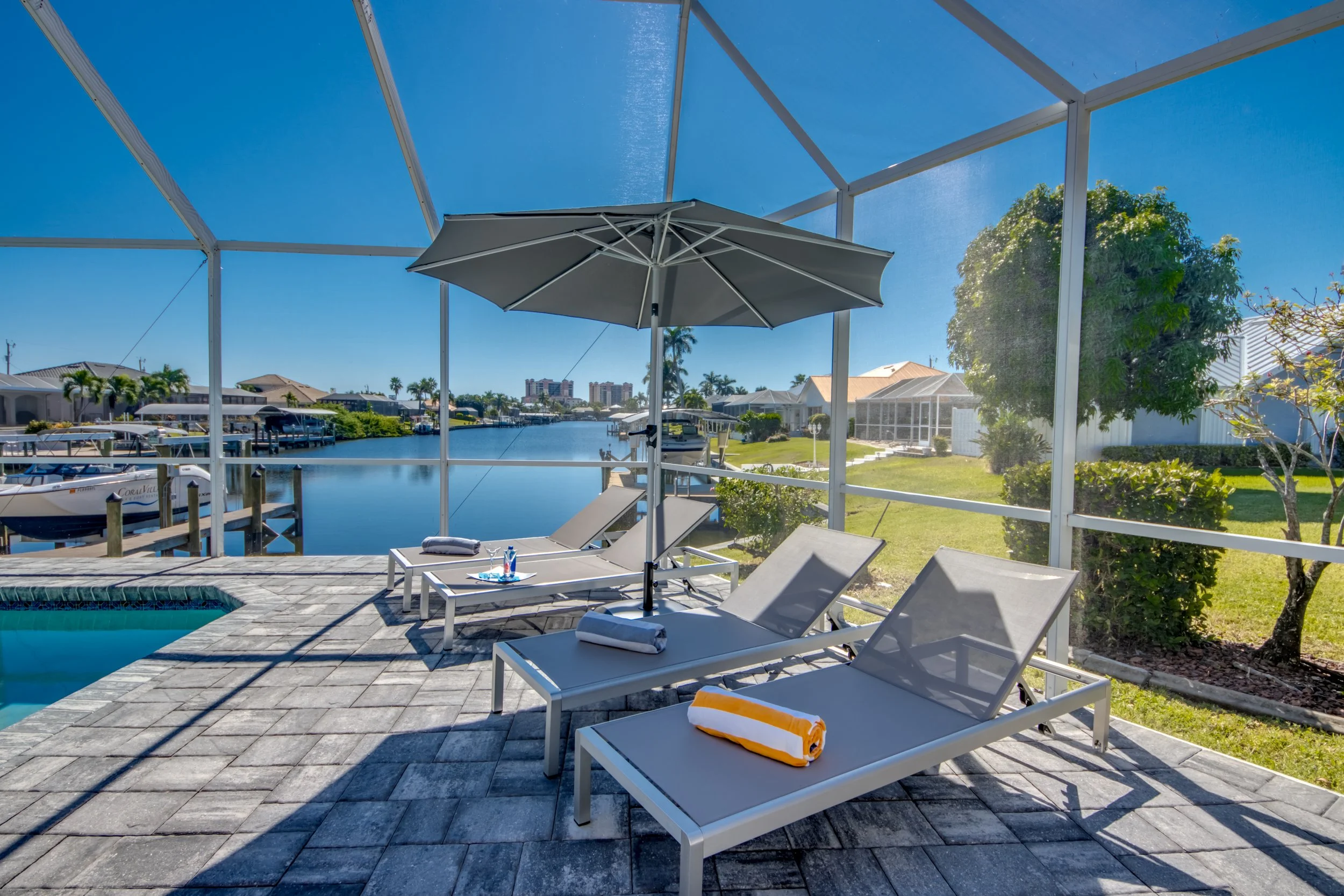 View of a screened-in patio area overlooking a canal with boats, houses, and trees. The patio has lounge chairs with towels and an umbrella, and a small pool in the foreground.