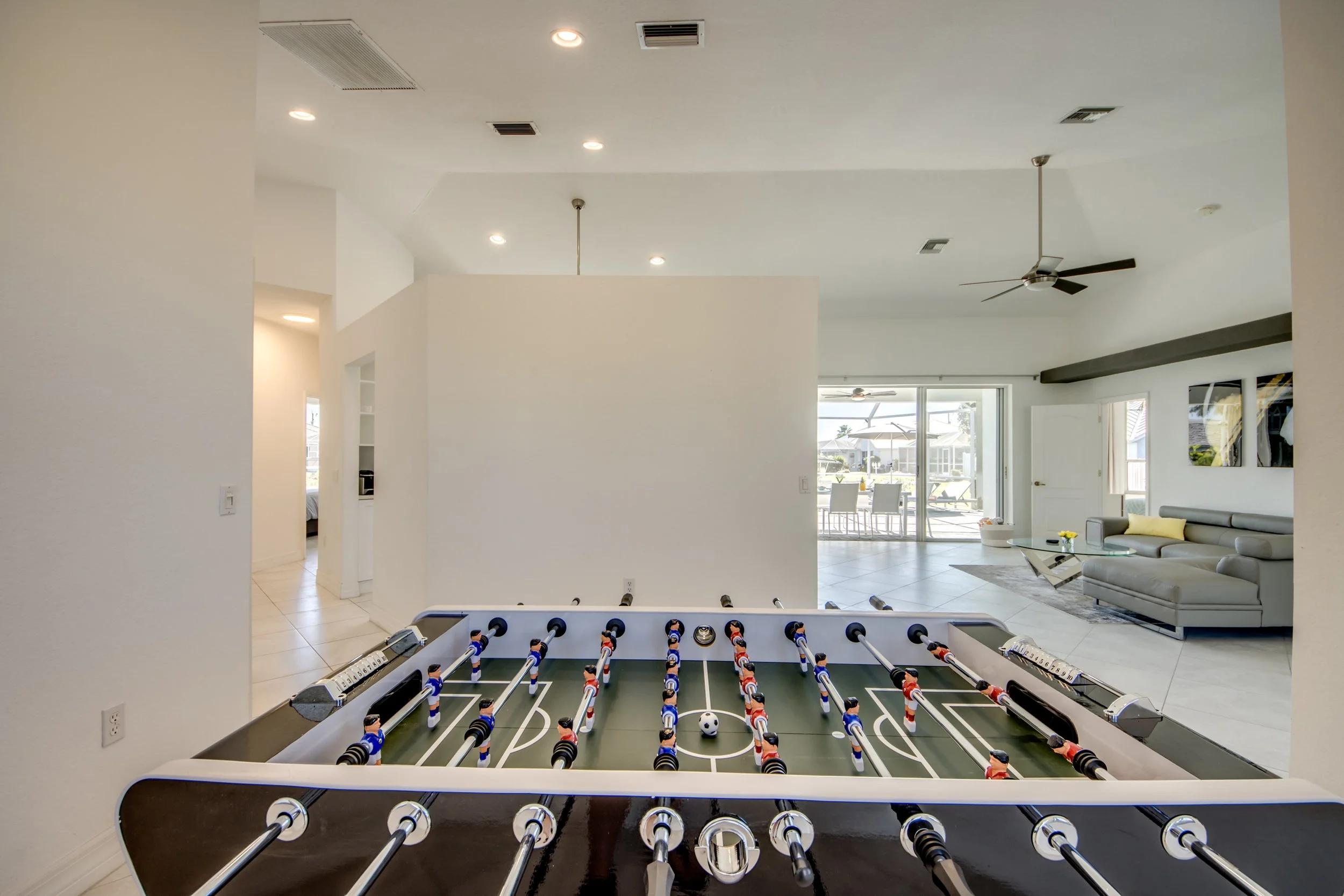 A modern open-concept living room with a foosball table in the foreground, leather sofa, a glass coffee table, and large sliding glass doors leading to a patio with chairs and umbrellas. Ceiling fans and recessed lighting are visible in the ceiling.