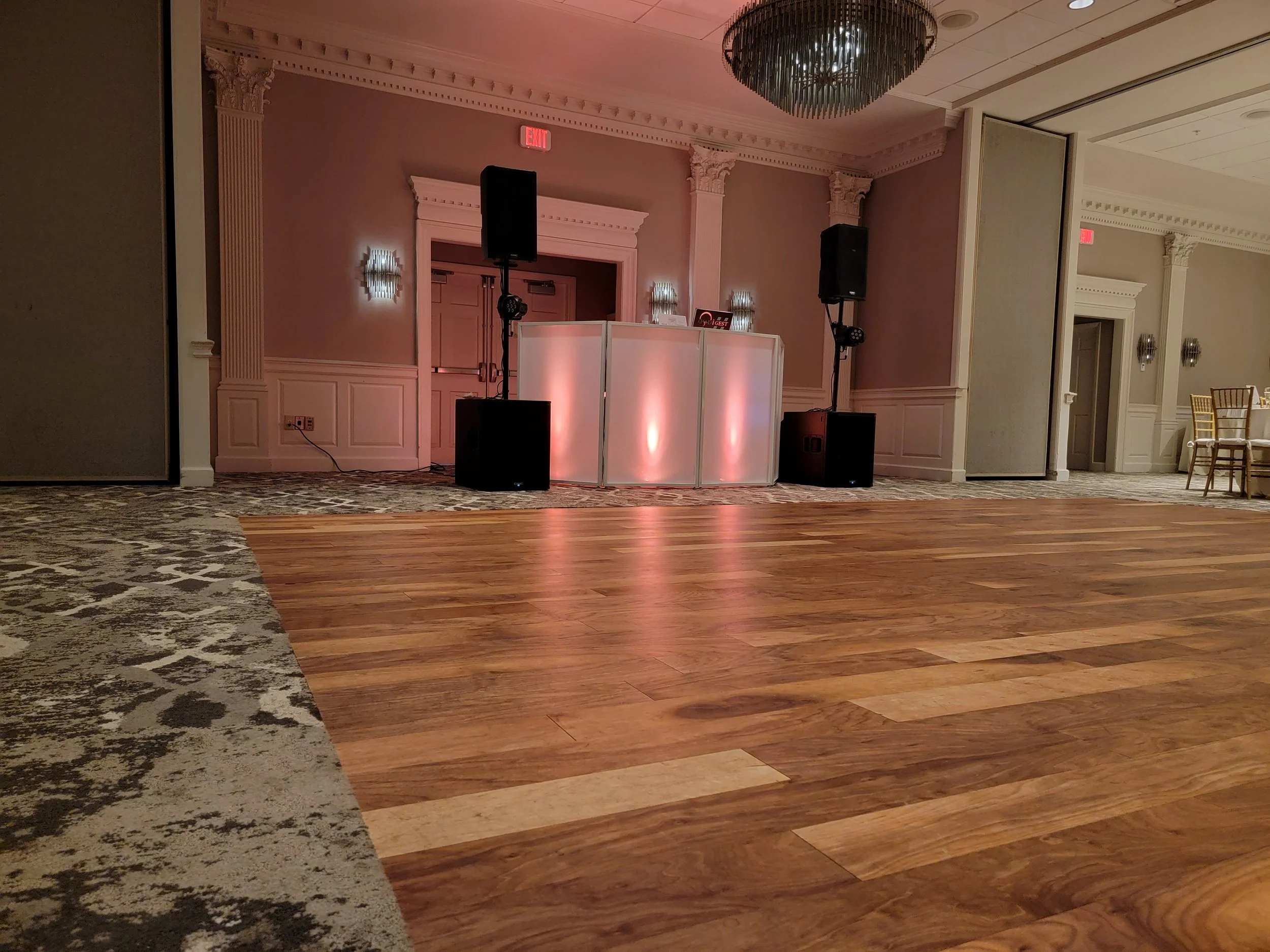 An indoor event space with a wooden dance floor, a DJ booth with lighting, large speakers, and wall-mounted lights, with chairs visible in the background.