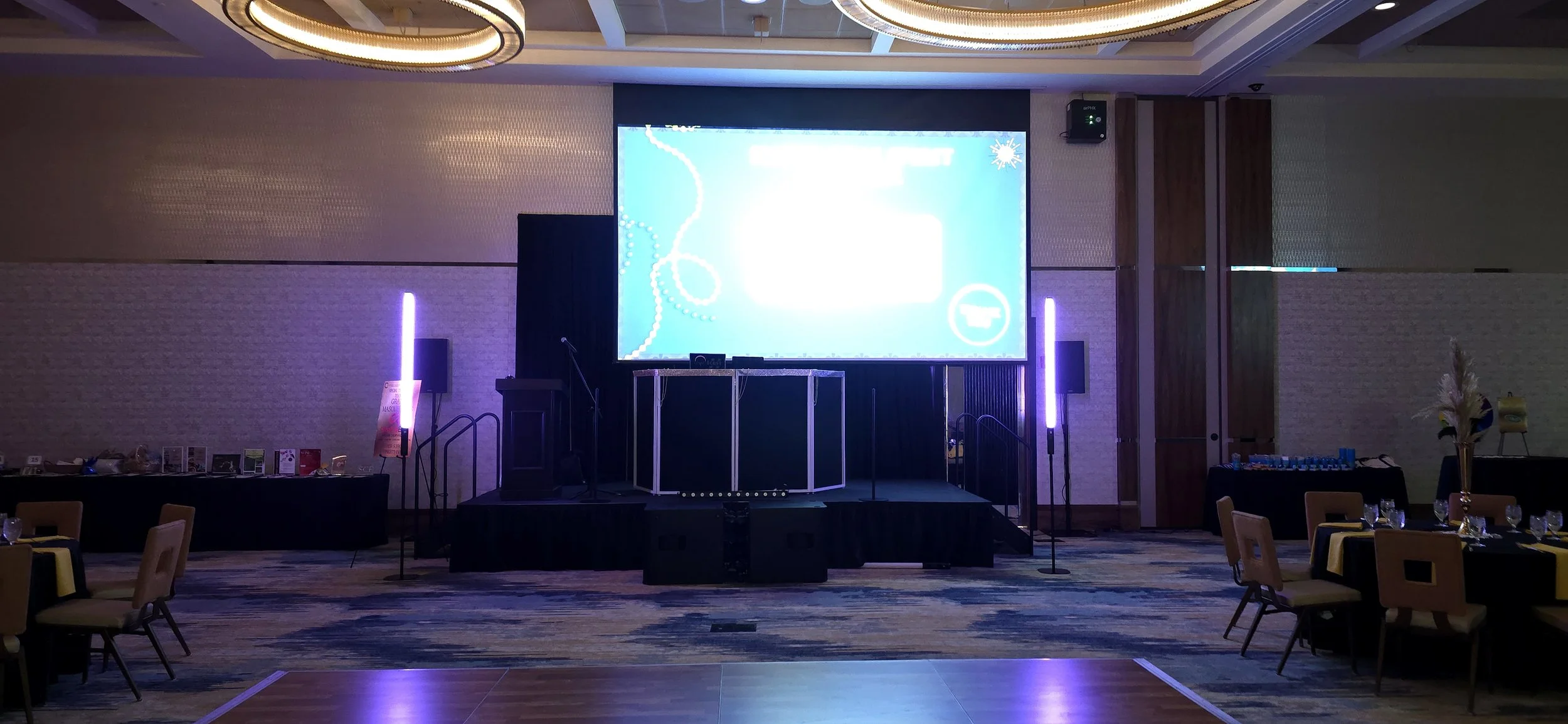 Empty event stage with a large illuminated screen, surrounded by a few purple LED light stands, with tables and chairs set up in front, and some decorations on the sides.