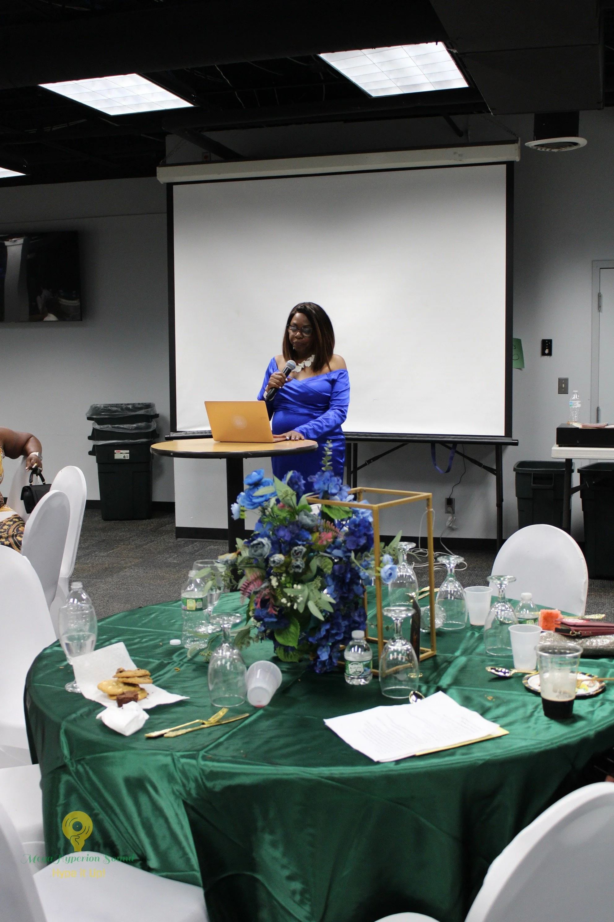 A woman in a blue dress speaking into a microphone, standing at a round table with a yellow laptop in front of her, in a conference room with a white projection screen behind her.