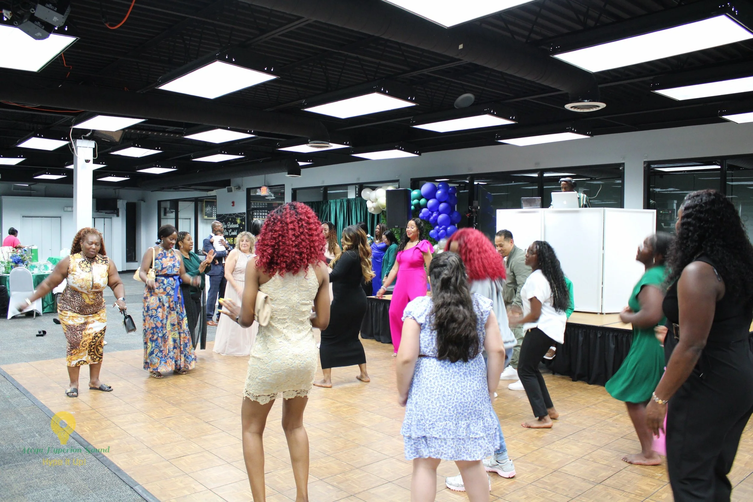 People dancing in a decorated indoor event space with balloons, a DJ setup, and a stage.