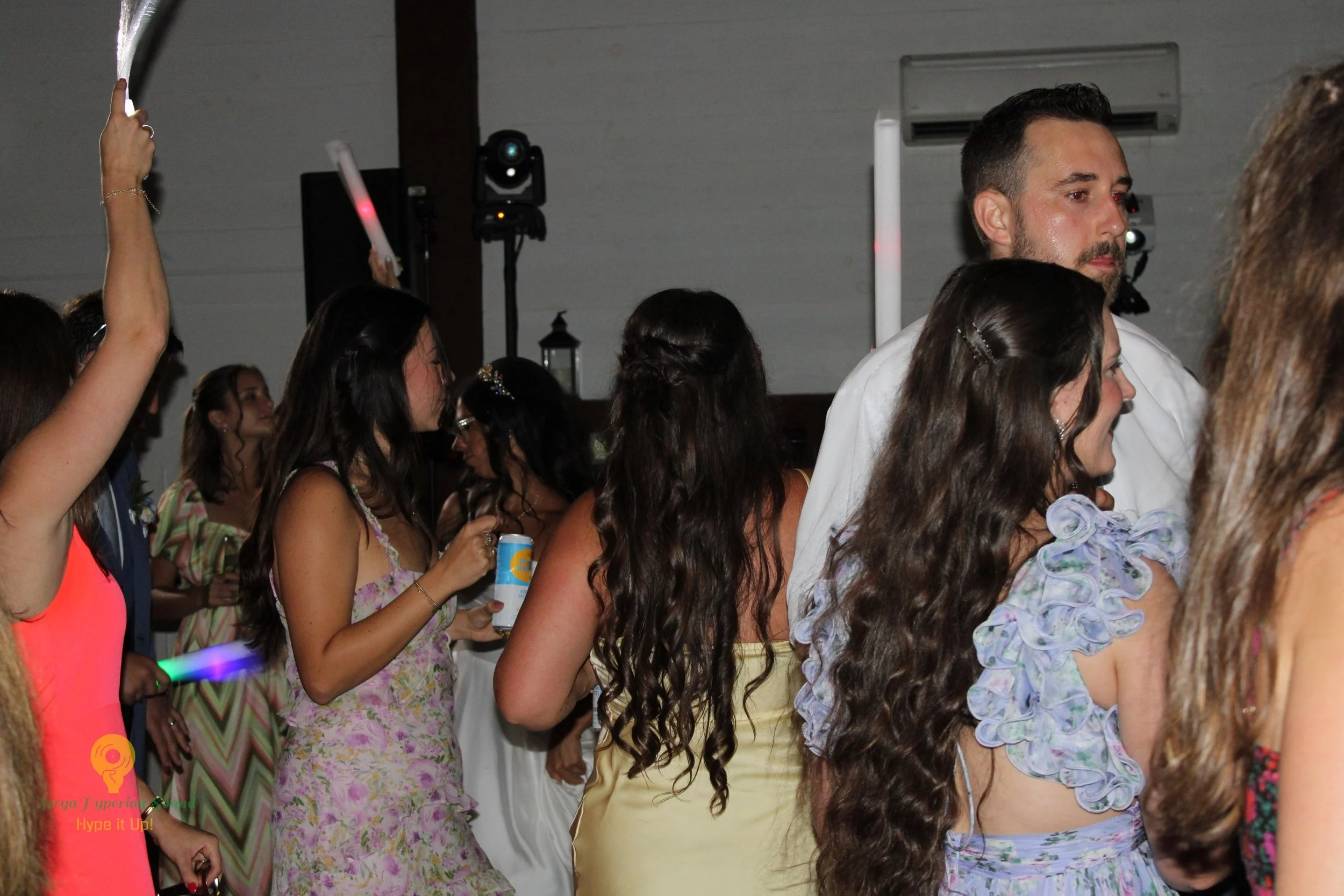 A group of people, mostly women, dressed in colorful attire with floral and patterned dresses, are standing closely together at an indoor event. Some are holding drinks and raising their hands or glow sticks. The setting appears festive and lively.