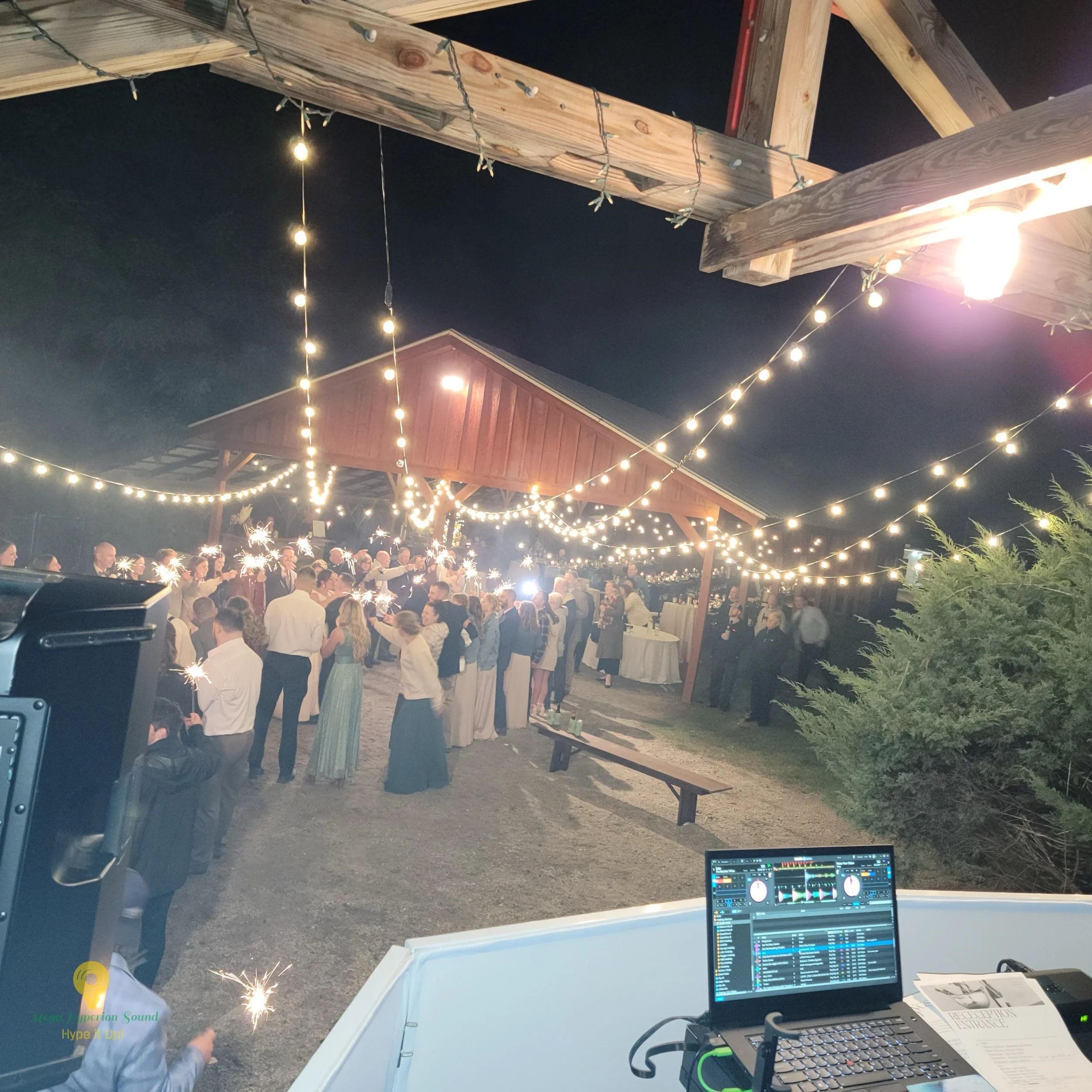 A party scene at night with string lights hanging from a wooden outdoor structure, a crowd of people holding sparklers, and a DJ booth with a laptop and sound equipment in the foreground.