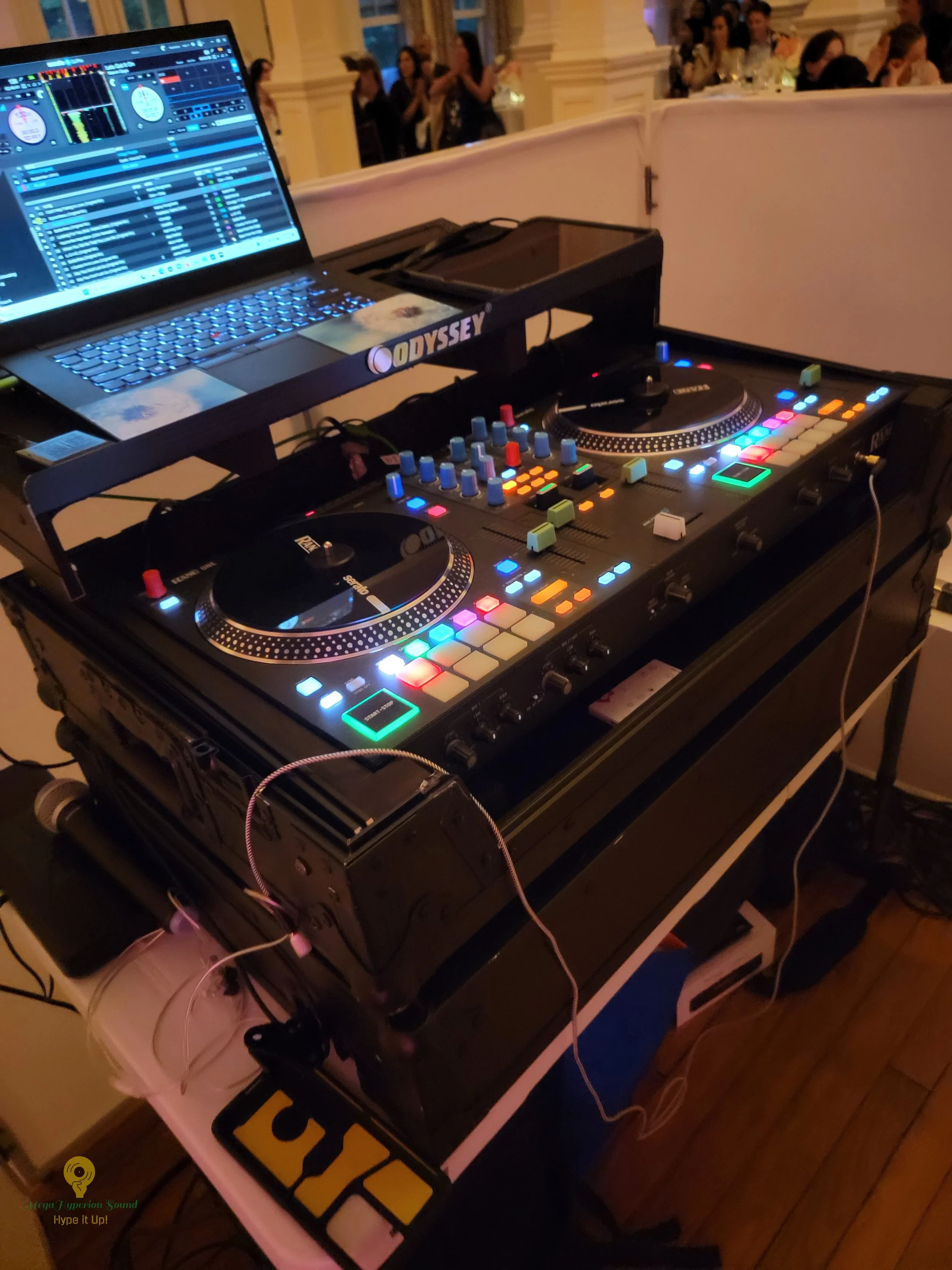 DJ equipment set up with a laptop and a DJ controller on a table, with people socializing in the background.