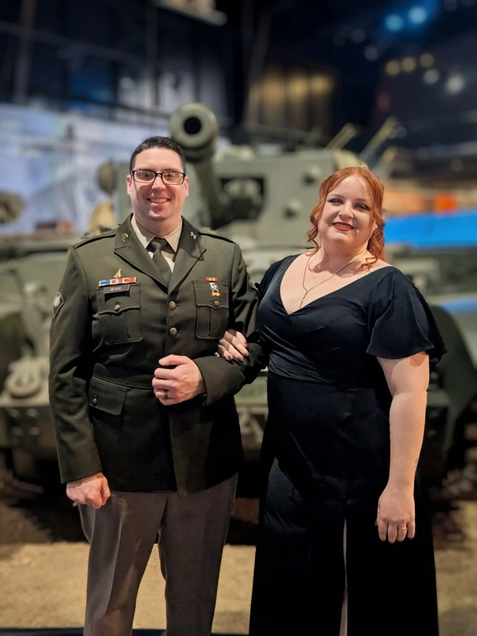 A man in military uniform and a woman in a black dress standing together in front of a military tank.