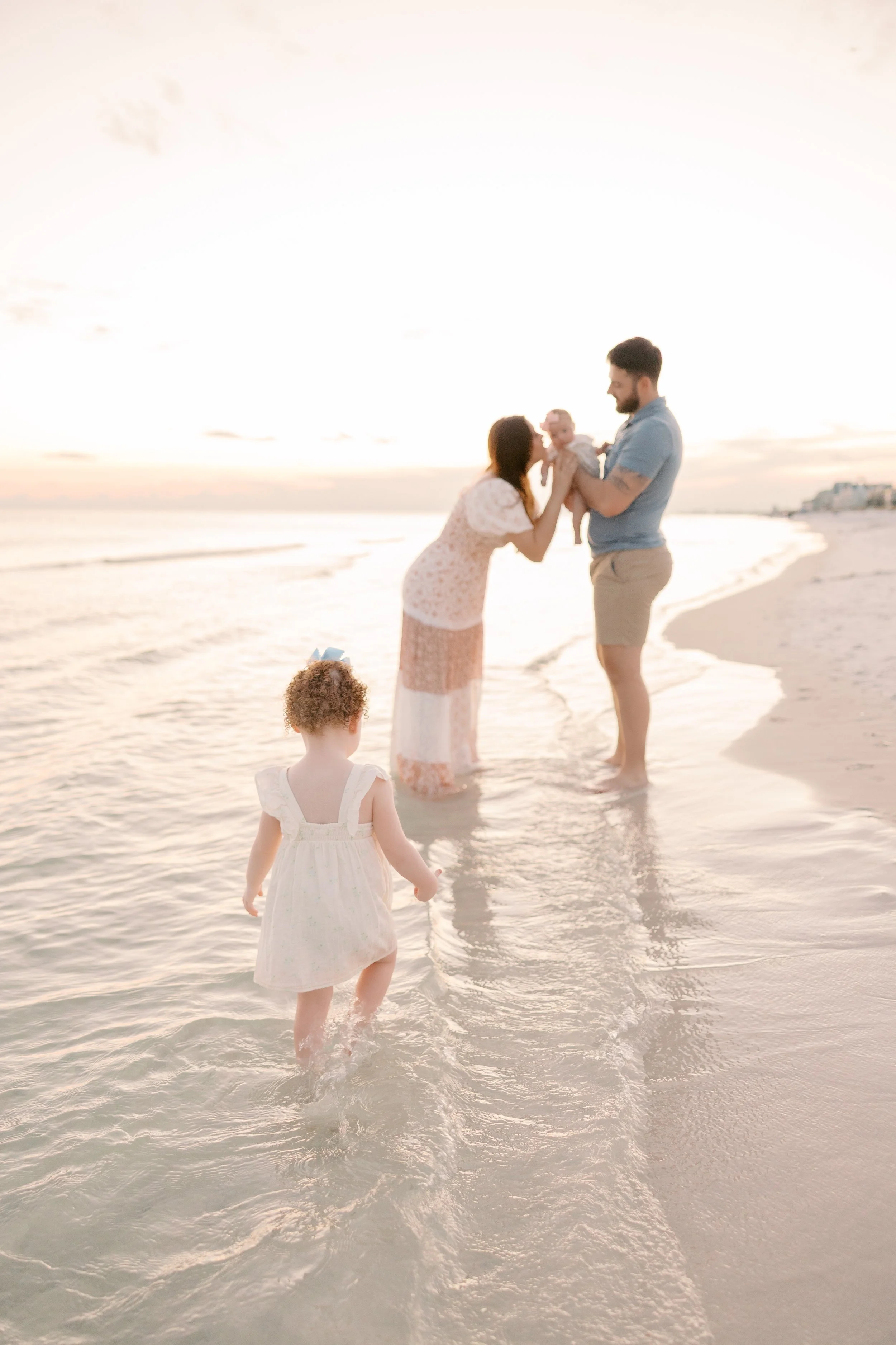 A family enjoying a walk on the beach at sunset, with two children and two adults near the shoreline, and the ocean and sky in the background.