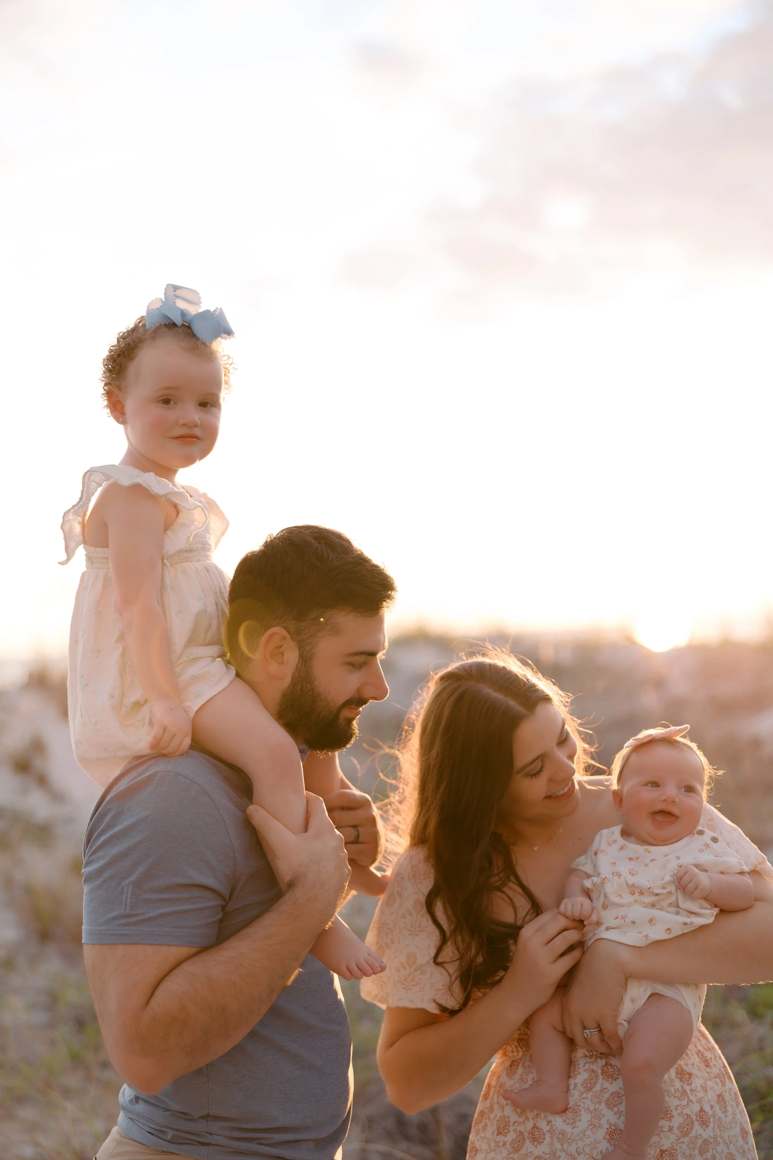 A family of four outdoors during sunset, with a man carrying a young girl on his shoulders, a woman holding a baby, and the sunlight illuminating their faces.
