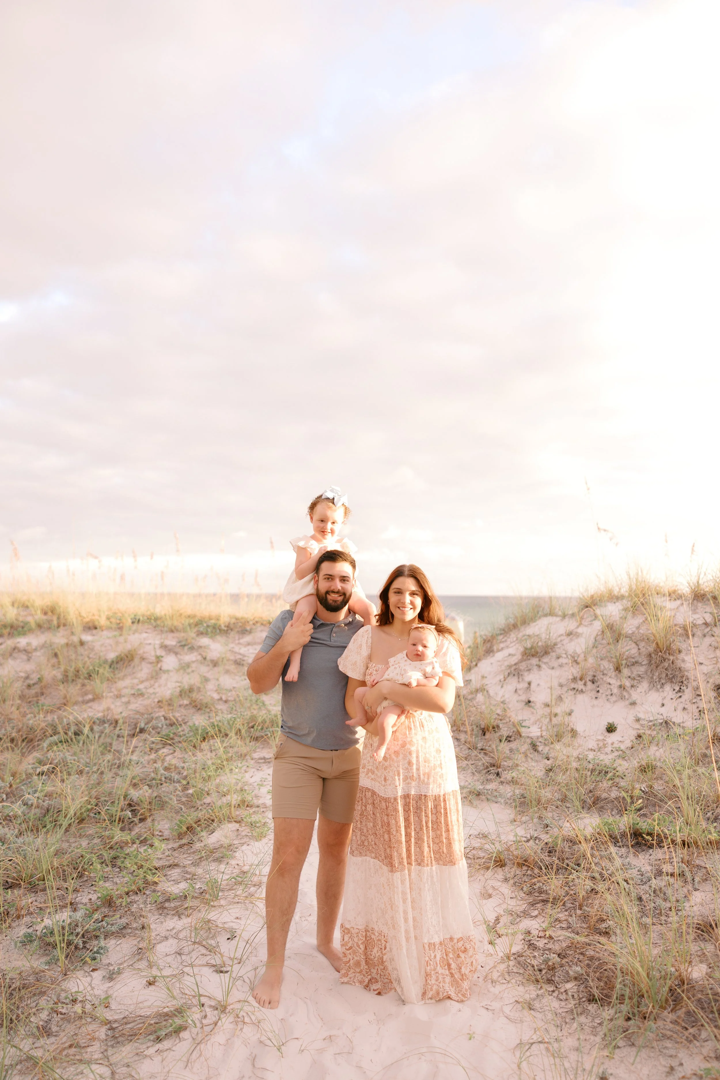 A family of four standing on a sandy beach with grassy dunes, under a cloud-filled sky, smiling at the camera.