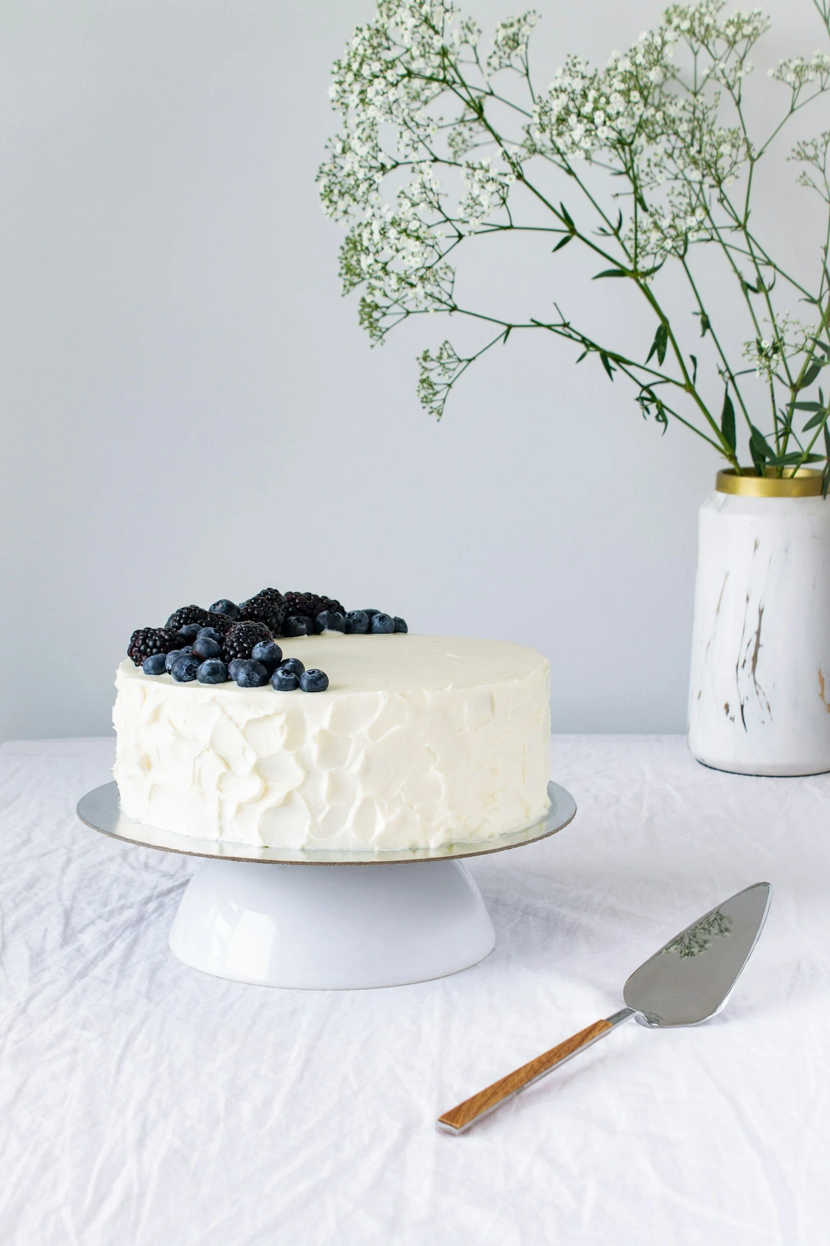 A white frosted cake decorated with blackberries and blueberries on top, placed on a white cake stand on a white tablecloth. In the background, there is a white vase with gold accents and green foliage flowers. A cake server with a wooden handle is positioned in front of the cake.
