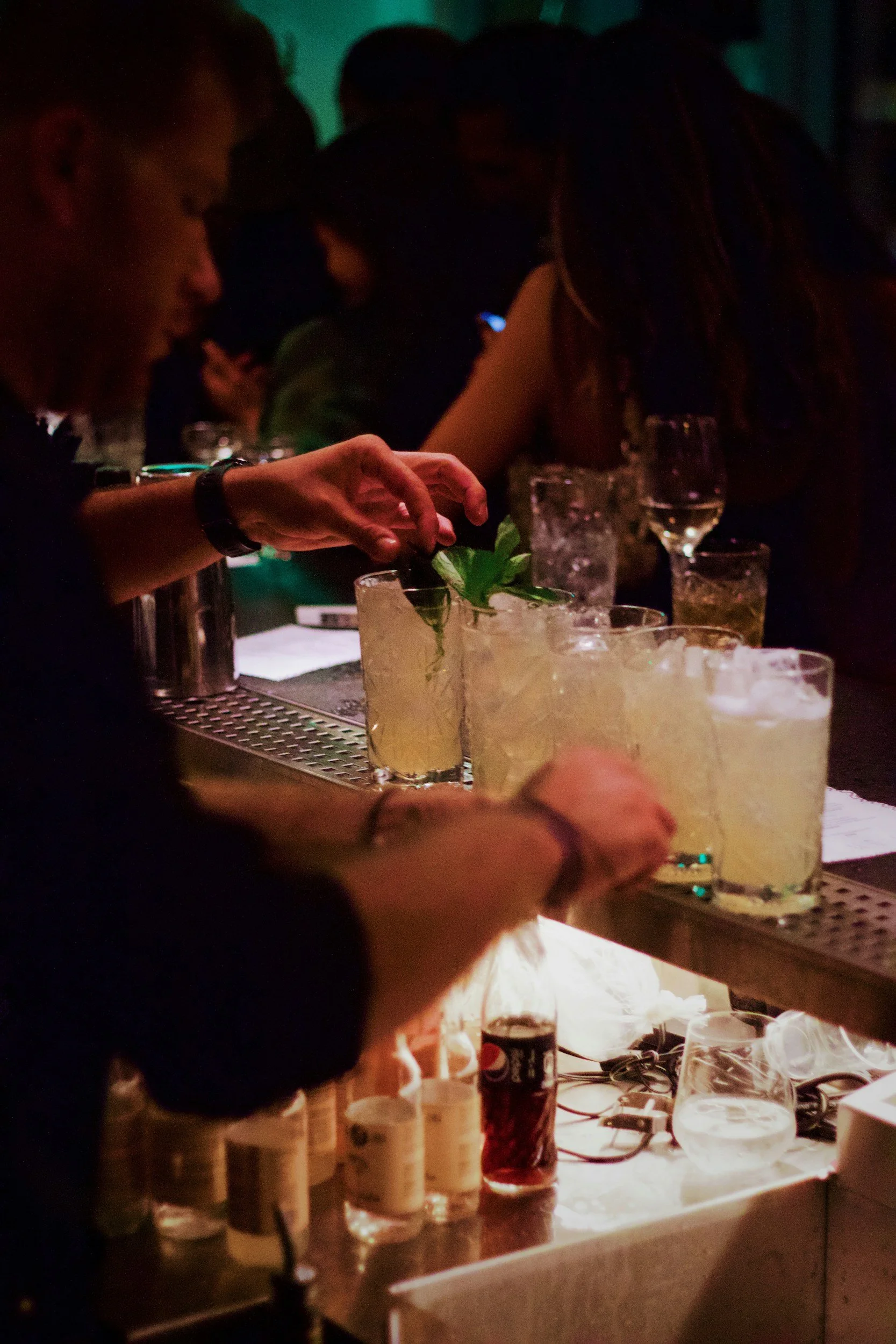 People at a bar or restaurant preparing cocktails or drinks with various bottles and ingredients on the counter, dim lighting background.