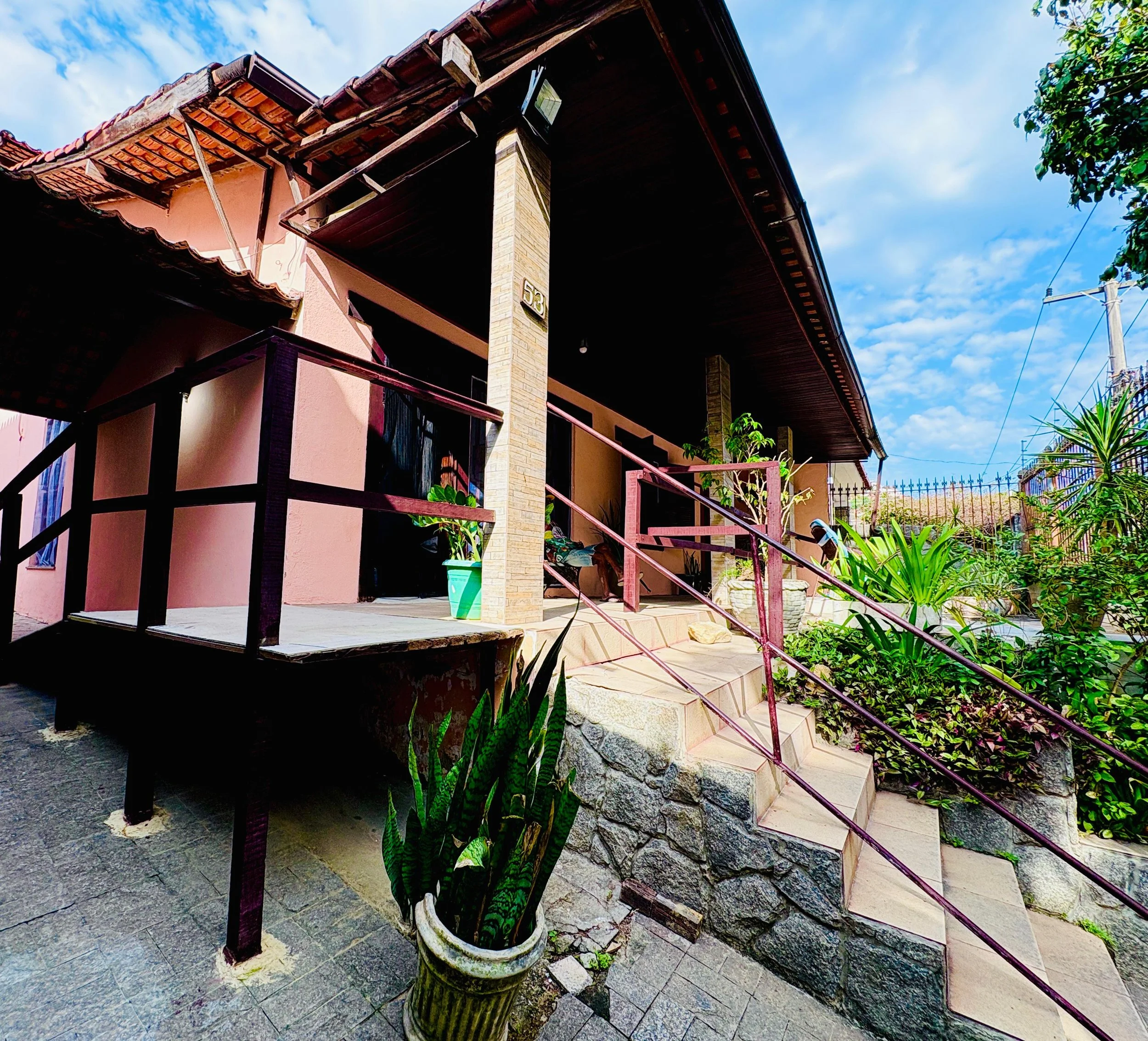 Casa de cor rosa com varanda e escadaria, jardim com plantas verdes, vaso com planta grande, parede de tijolos, céu com nuvens, poste de energia elétrica ao lado.