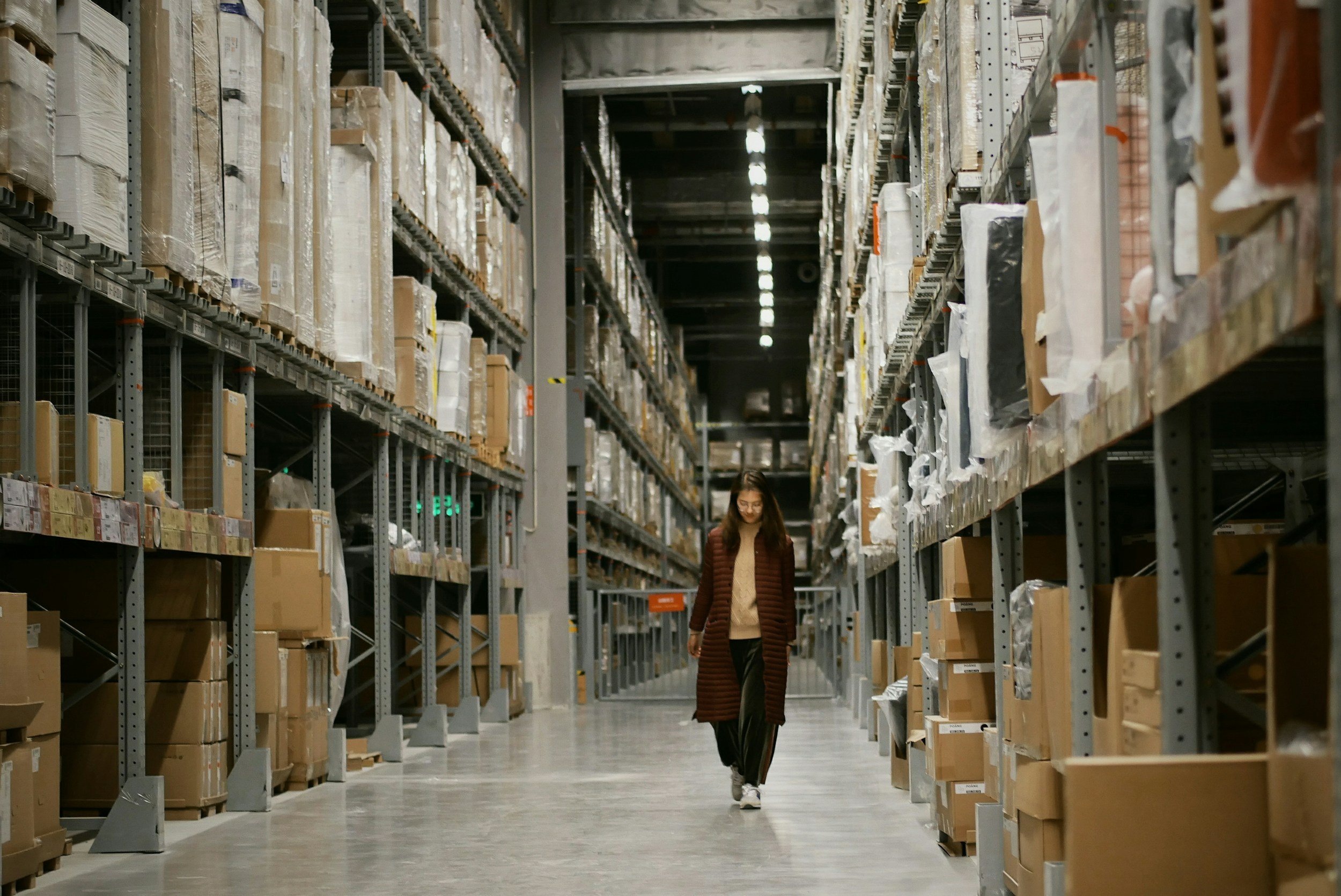 A woman walking through an aisle filled with shelves of brown boxes in a large warehouse.