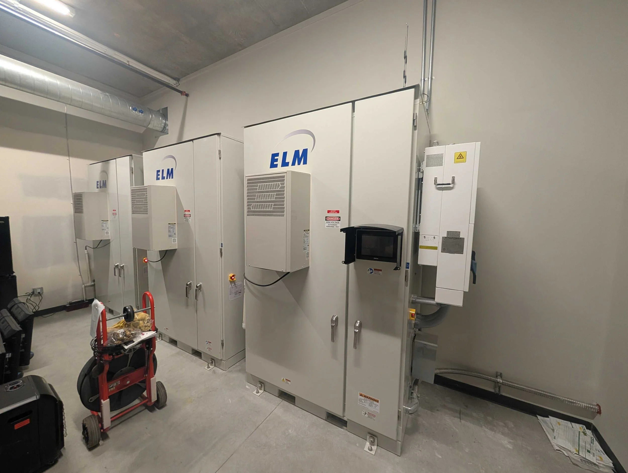A row of industrial electrical panels with the logo ELM on each, mounted on a wall in a utility room, with a small red cart and some tools in front.