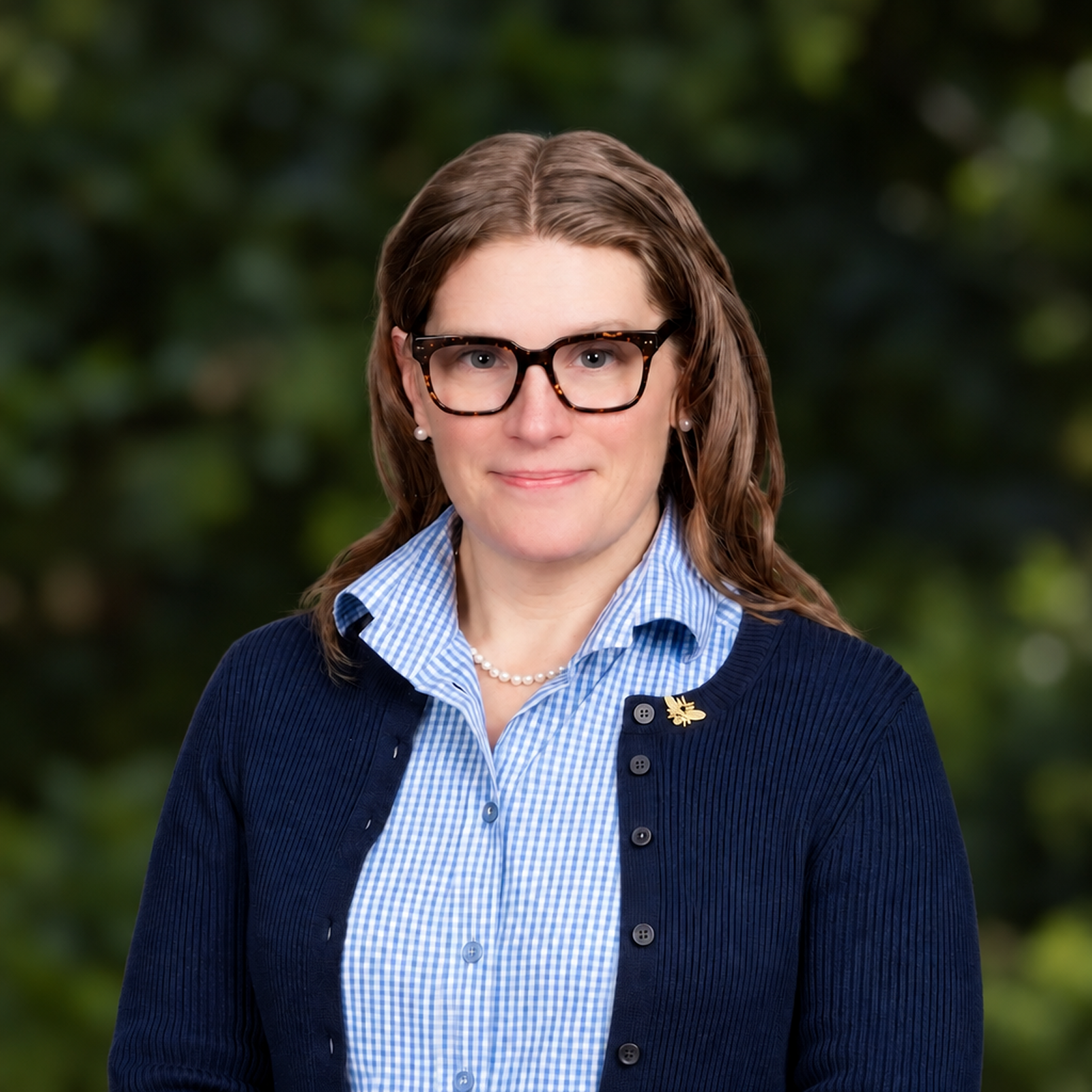 A woman with brown hair, glasses, pearl earrings, and a pearl necklace wearing a blue checkered shirt and a navy cardigan with a gold pin, standing outdoors with a blurry green background.