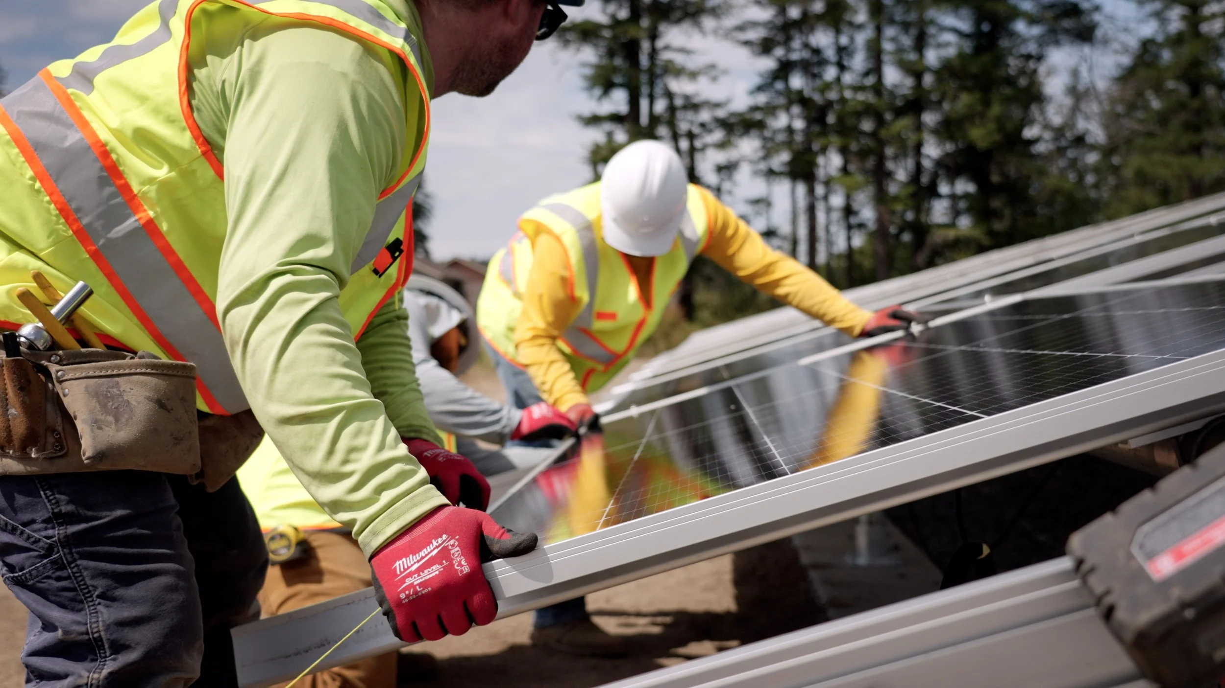 Two workers in yellow safety vests and helmets installing solar panels outdoors, with trees and cloudy sky in the background.