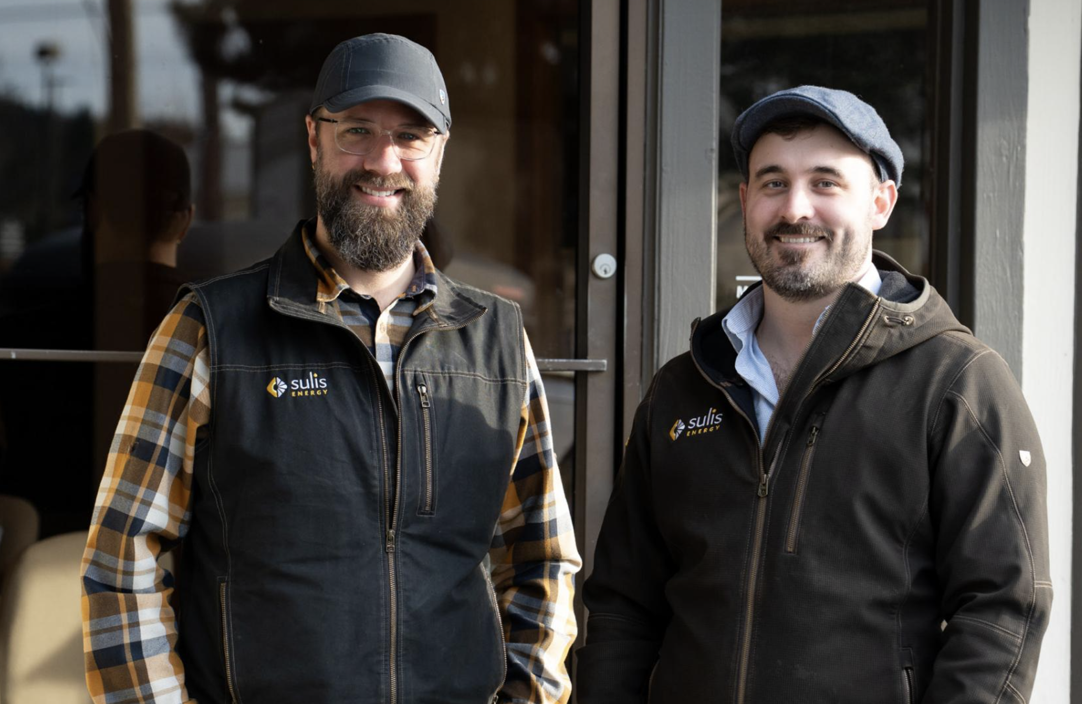 Two smiling men standing outside, wearing black vests with the logo 'Sulis Energy'. One man has a beard, glasses, and a gray cap, while the other has a beard and is wearing a gray flat cap.