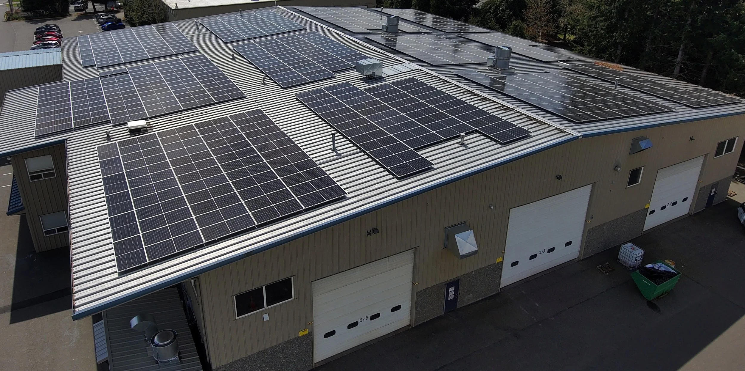 Aerial view of a building with a metal roof covered with multiple solar panels and rooftop equipment, surrounded by trees, parking lot, and other structures.