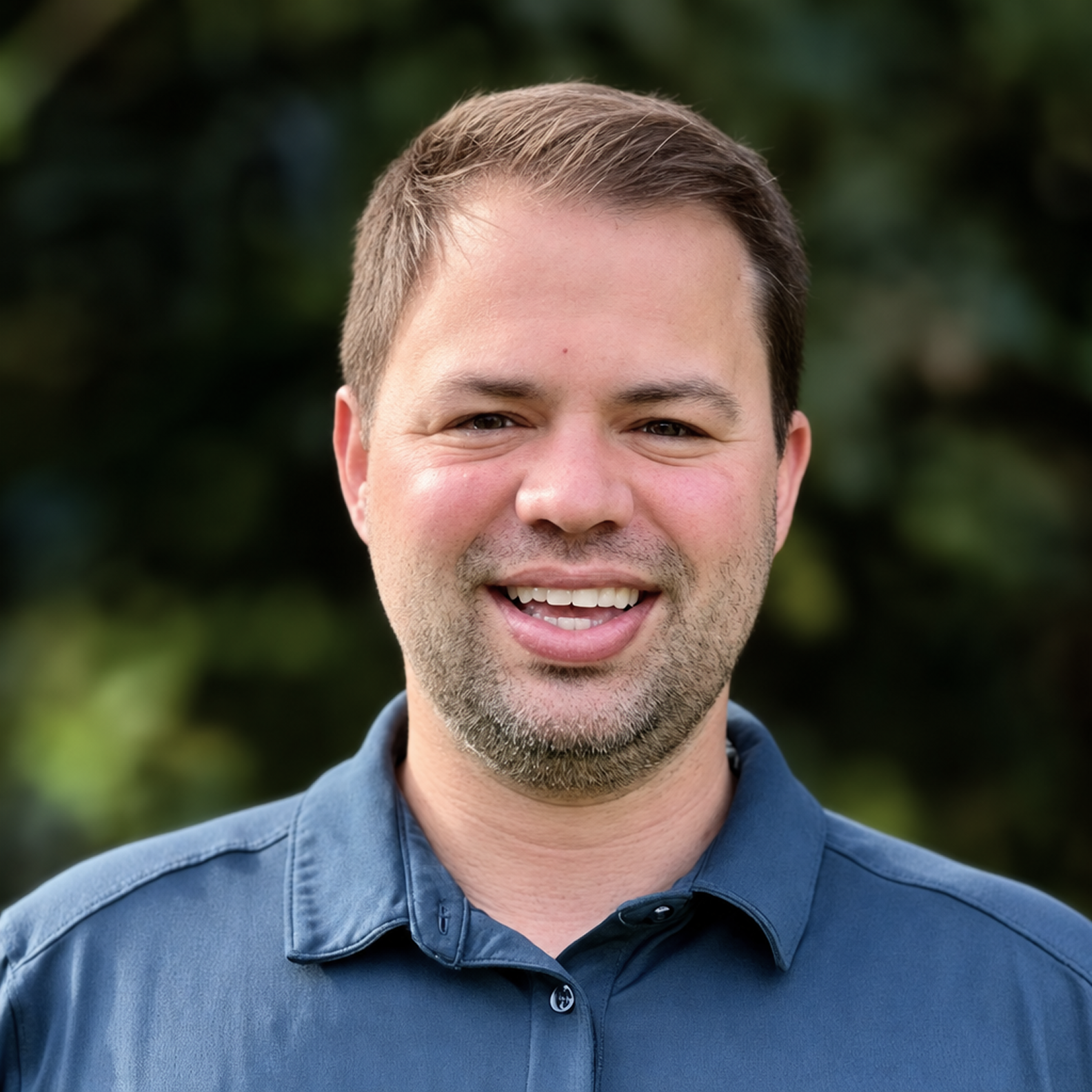 Close-up of a smiling man with short brown hair and a beard, wearing a navy blue collared shirt, outdoors with green foliage in the background.