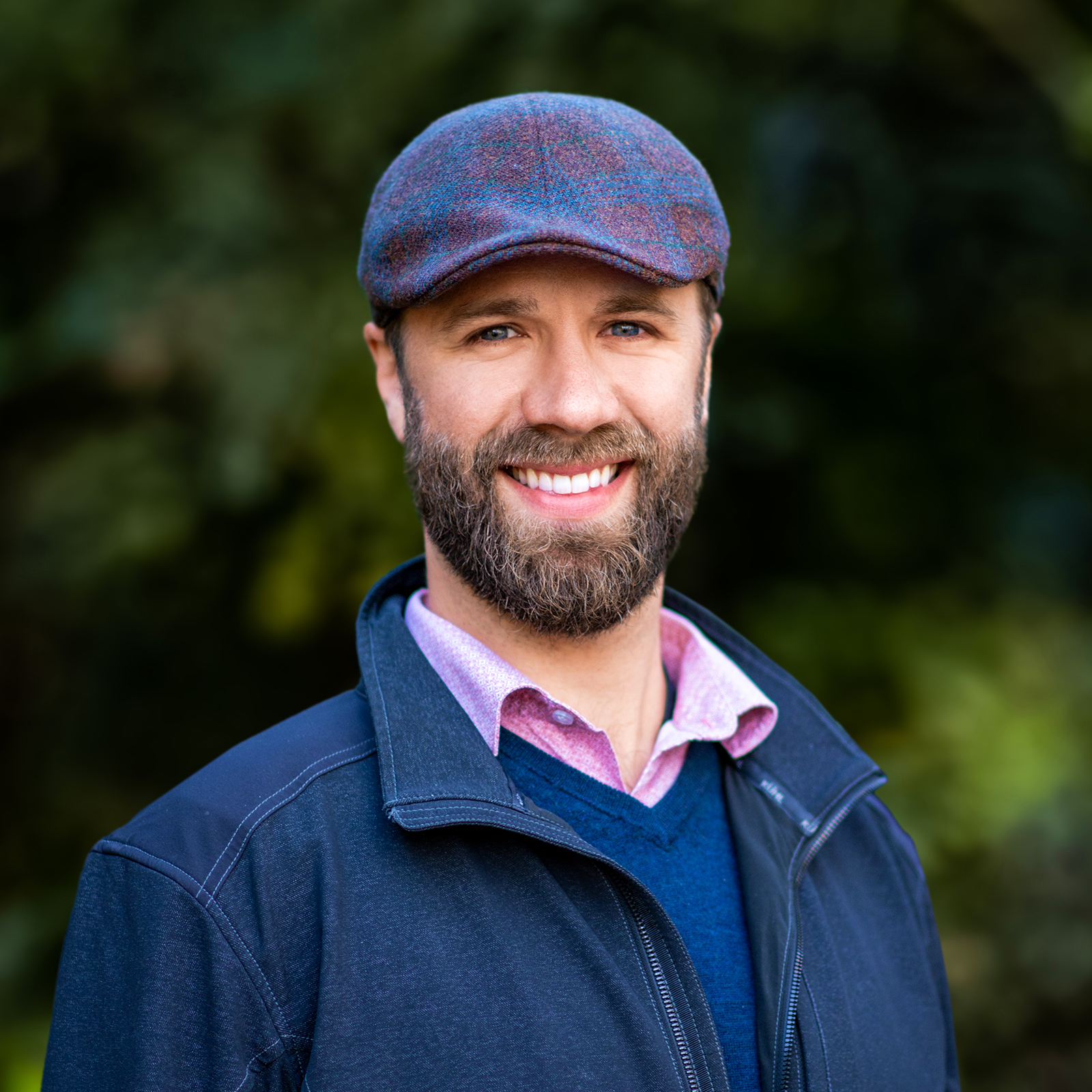 A smiling man with a beard wearing a blue flat cap, a pink shirt, and a navy jacket, standing outdoors with green foliage in the background.