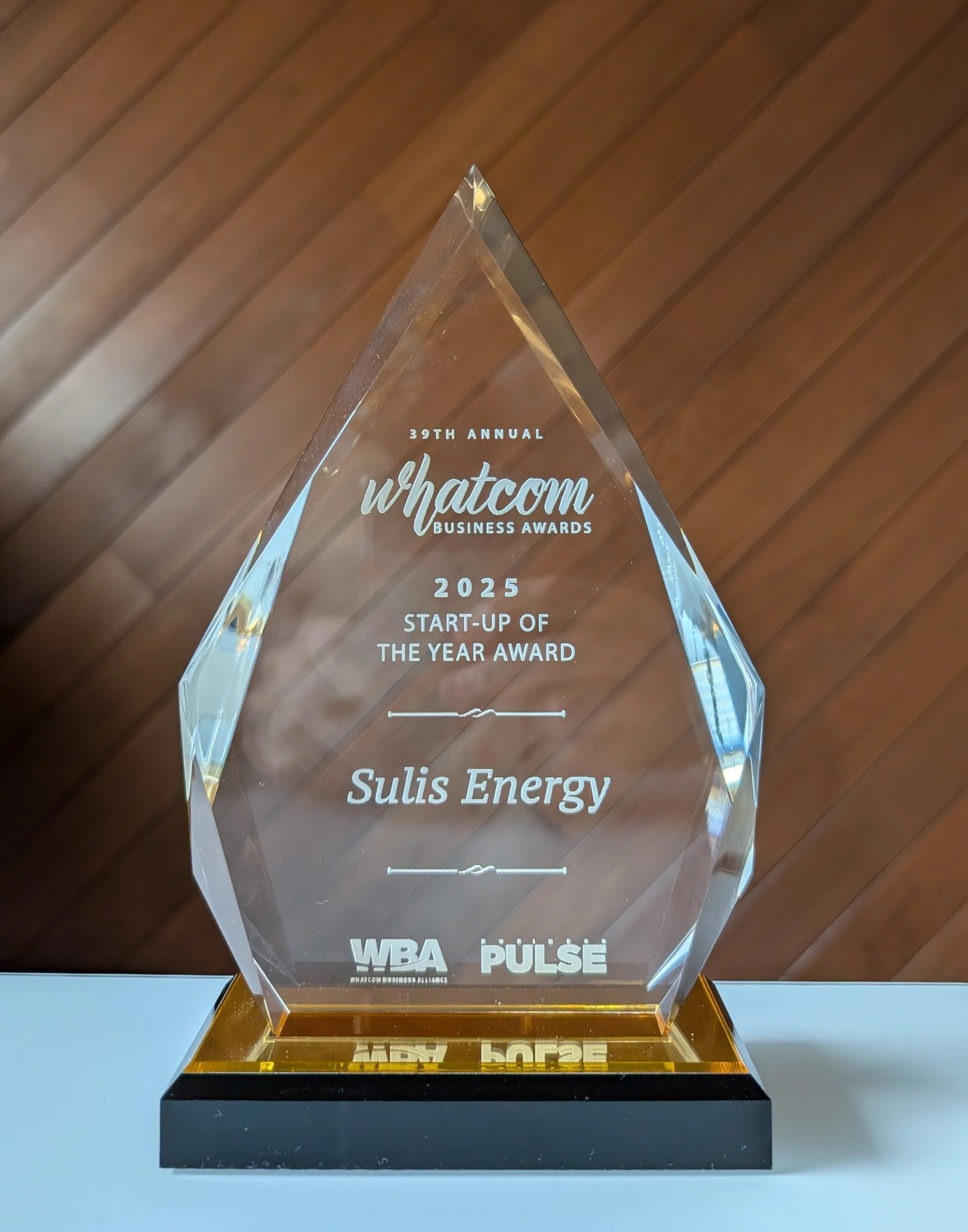Glass award plaque with inscribed text, set on a black and gold base, with a wooden background.