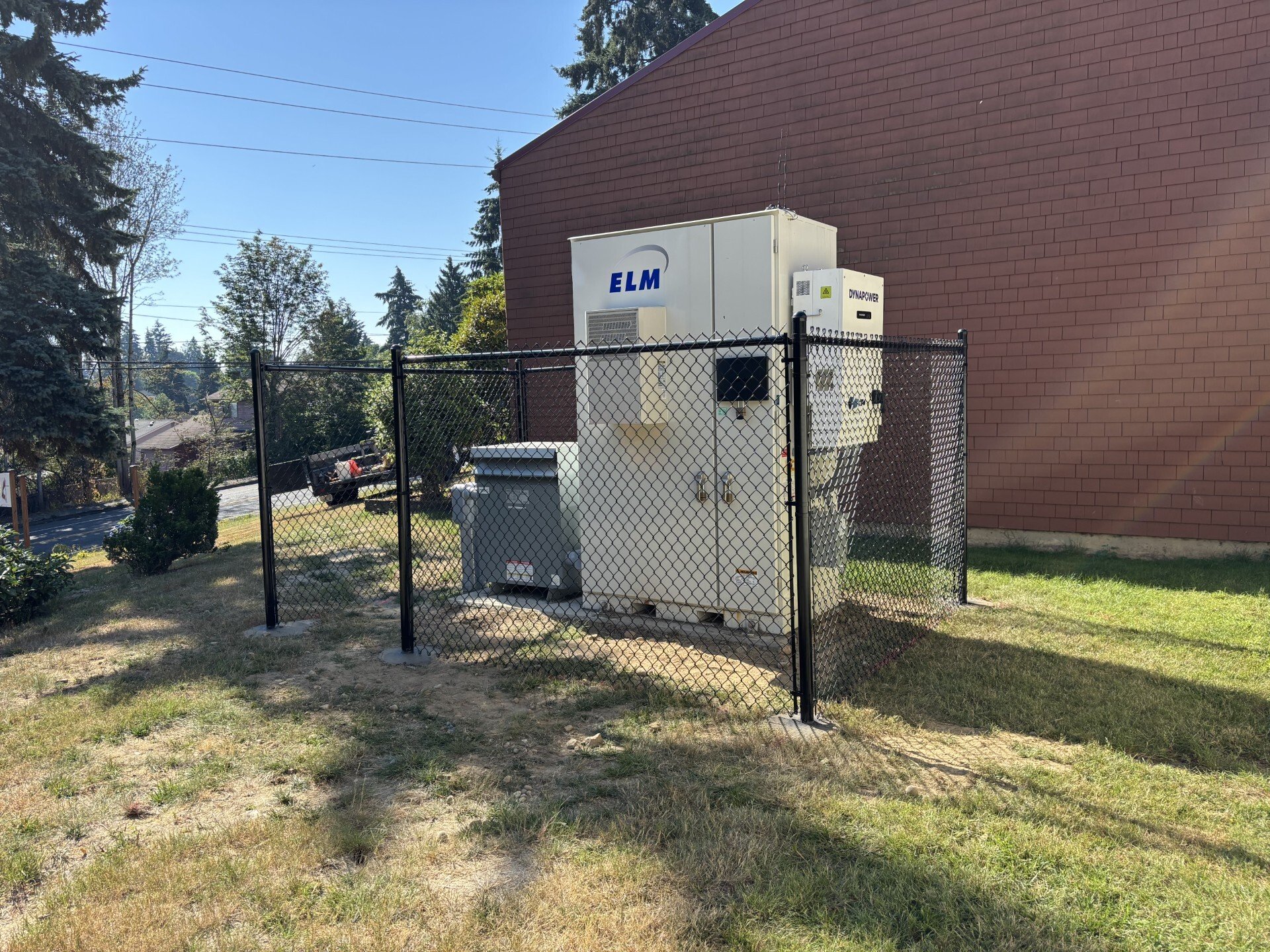 An electrical transformer and equipment enclosed within a black chain-link fence on a grassy yard, next to a red brick building, with trees and a blue sky in the background.