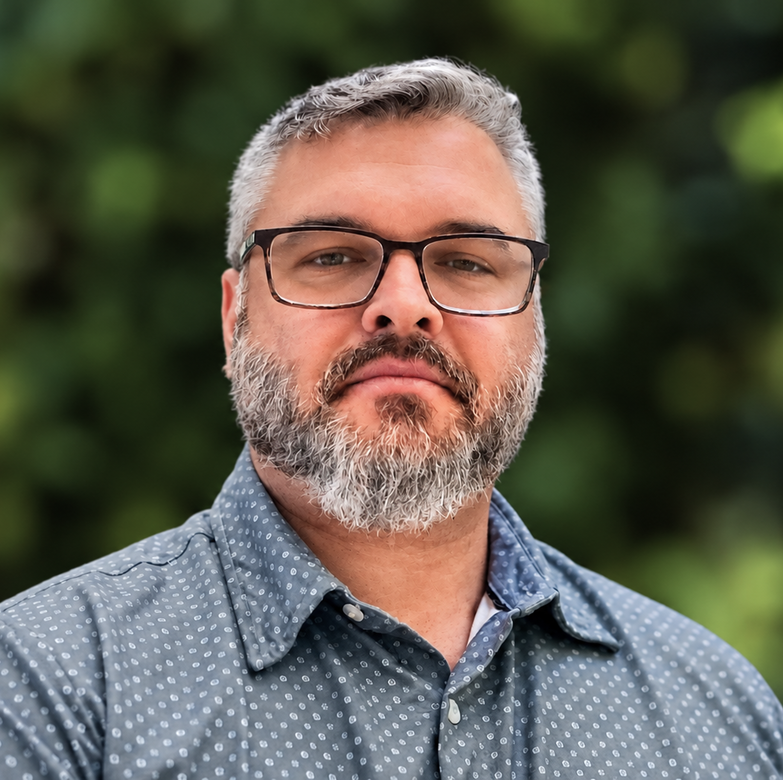 Close-up headshot of a middle-aged man with gray hair, a beard, and glasses wearing a blue patterned shirt outdoors with blurred greenery in the background.