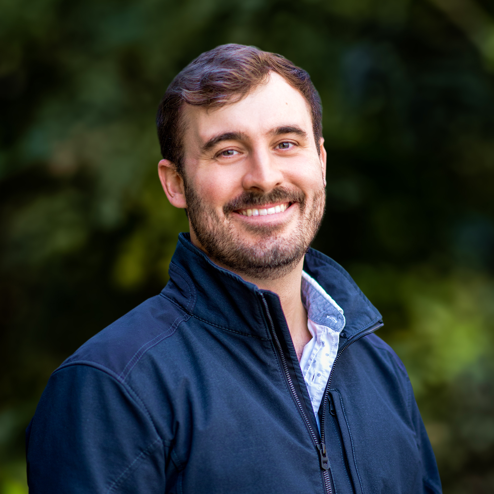 Headshot of a smiling man with brown hair and a beard, wearing a dark blue zip-up jacket, outdoors with a blurred green background.