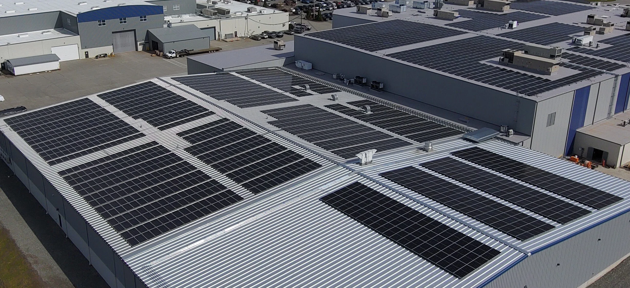 An aerial view of a large commercial building with a white metal roof covered in numerous solar panels. Surrounding buildings and parking areas are also visible.