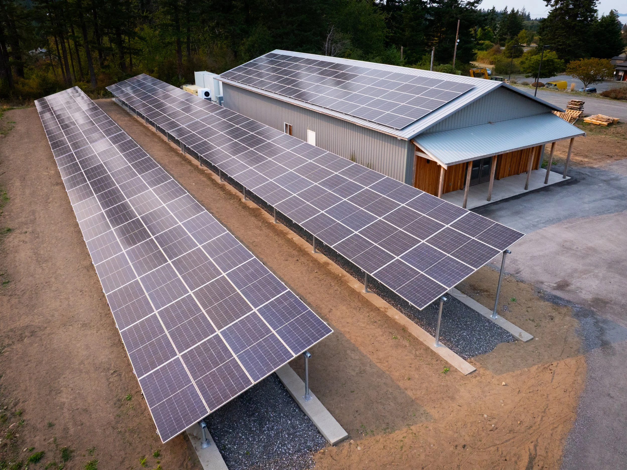 Building with solar panels on roof and on ground, surrounded by trees and a parking lot.