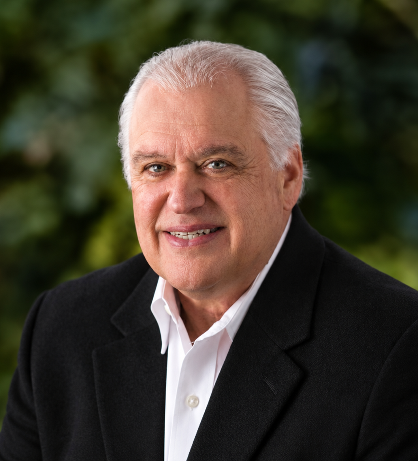 Portrait of an older man with white hair, wearing a black suit jacket and white shirt, smiling in front of a green blurred background.