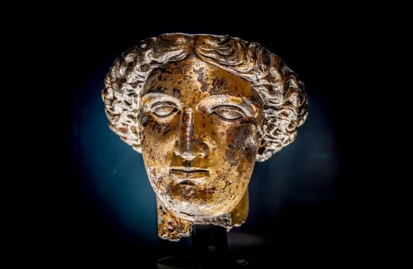 Ancient Greek or Roman marble sculpture bust of a woman with curly hair, showing signs of aging and damage, set against a dark background.