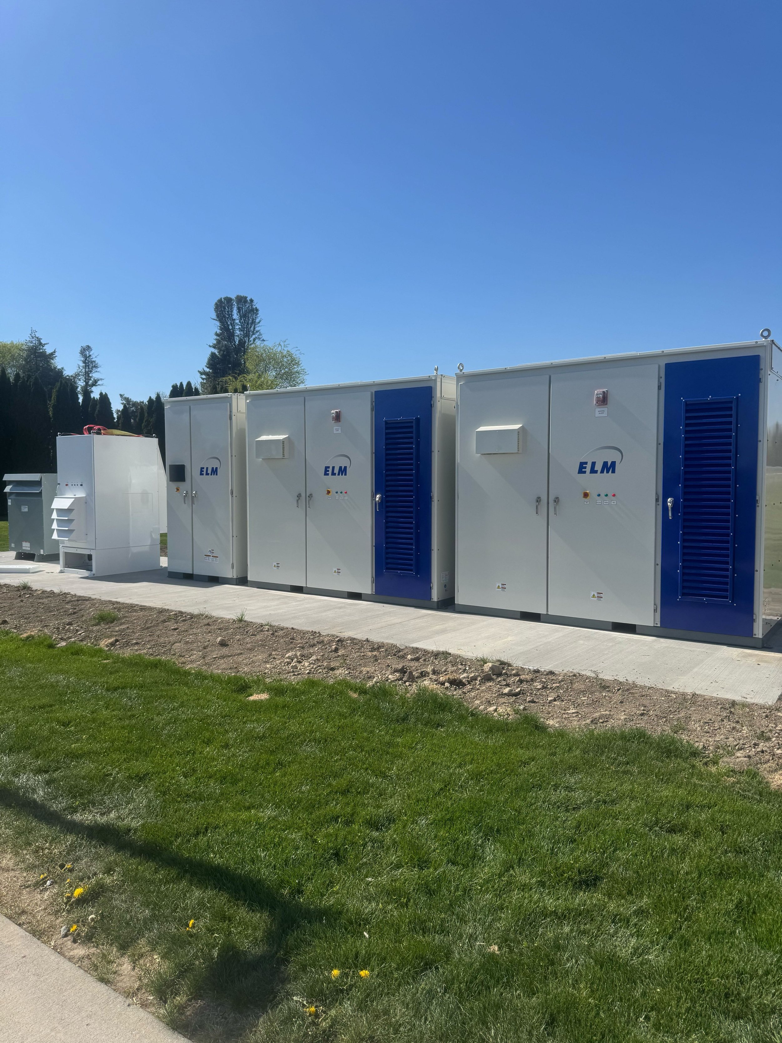 Outdoor electrical infrastructure with several large white and blue electrical cabinets, green grass in the foreground, and tall trees under a clear blue sky.