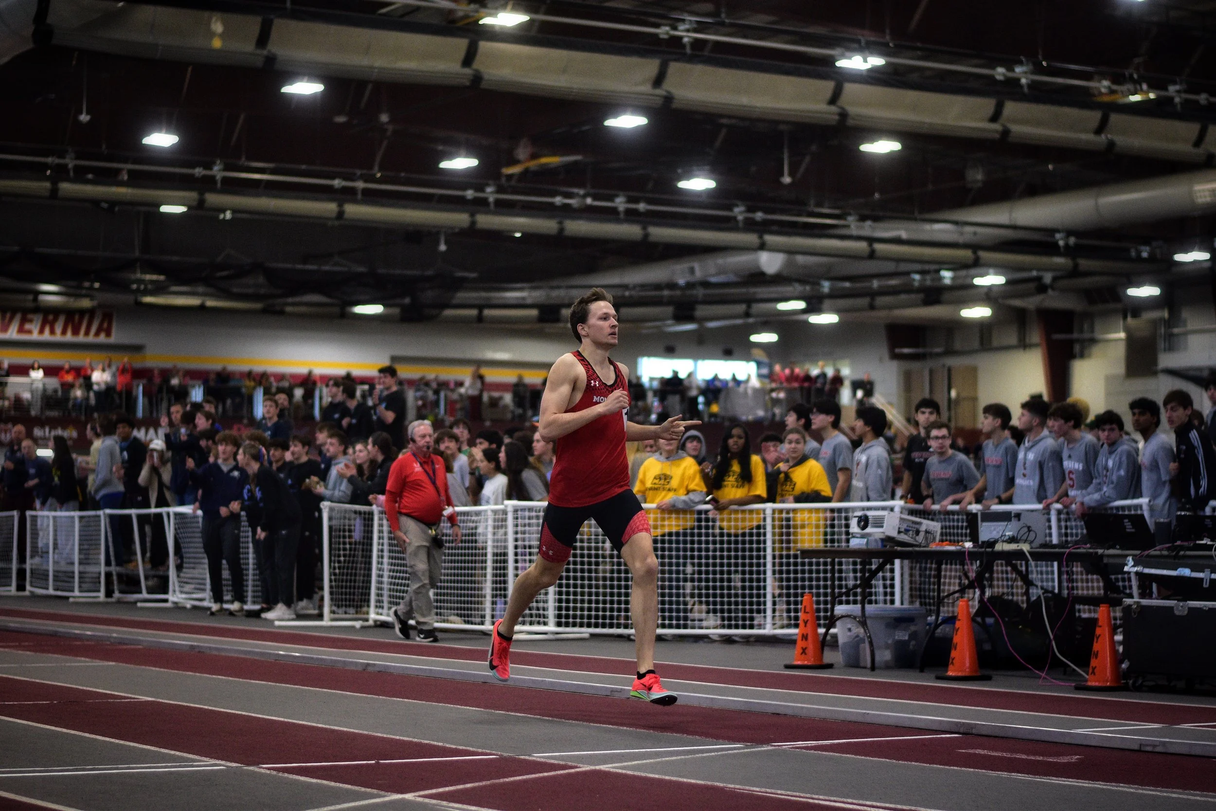 Male athlete in red running gear participating in an indoor track event with a crowd and officials watching.