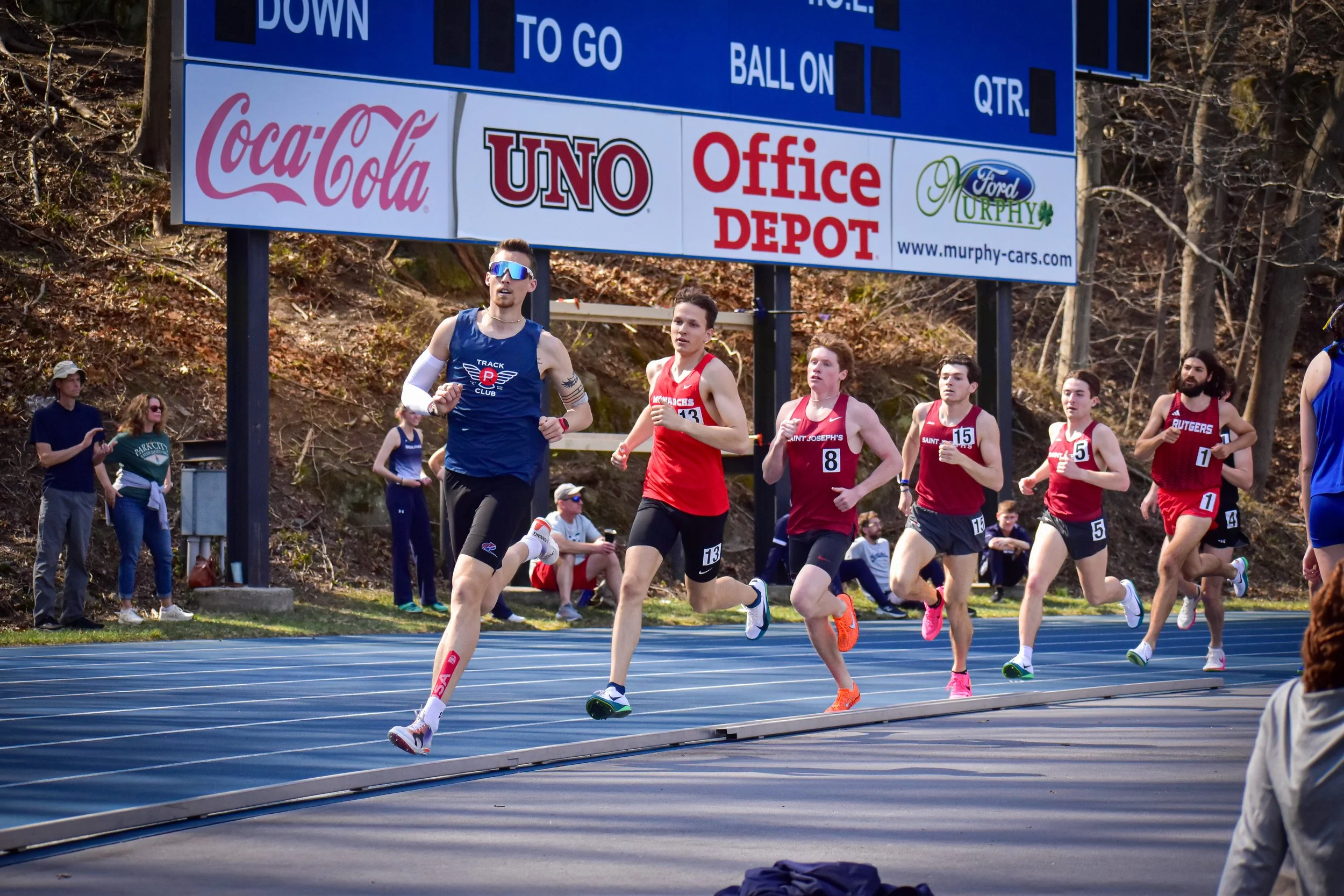 A group of runners competing in a track race with a blue track, surrounded by spectators and trees, and a large sponsor billboard overhead.