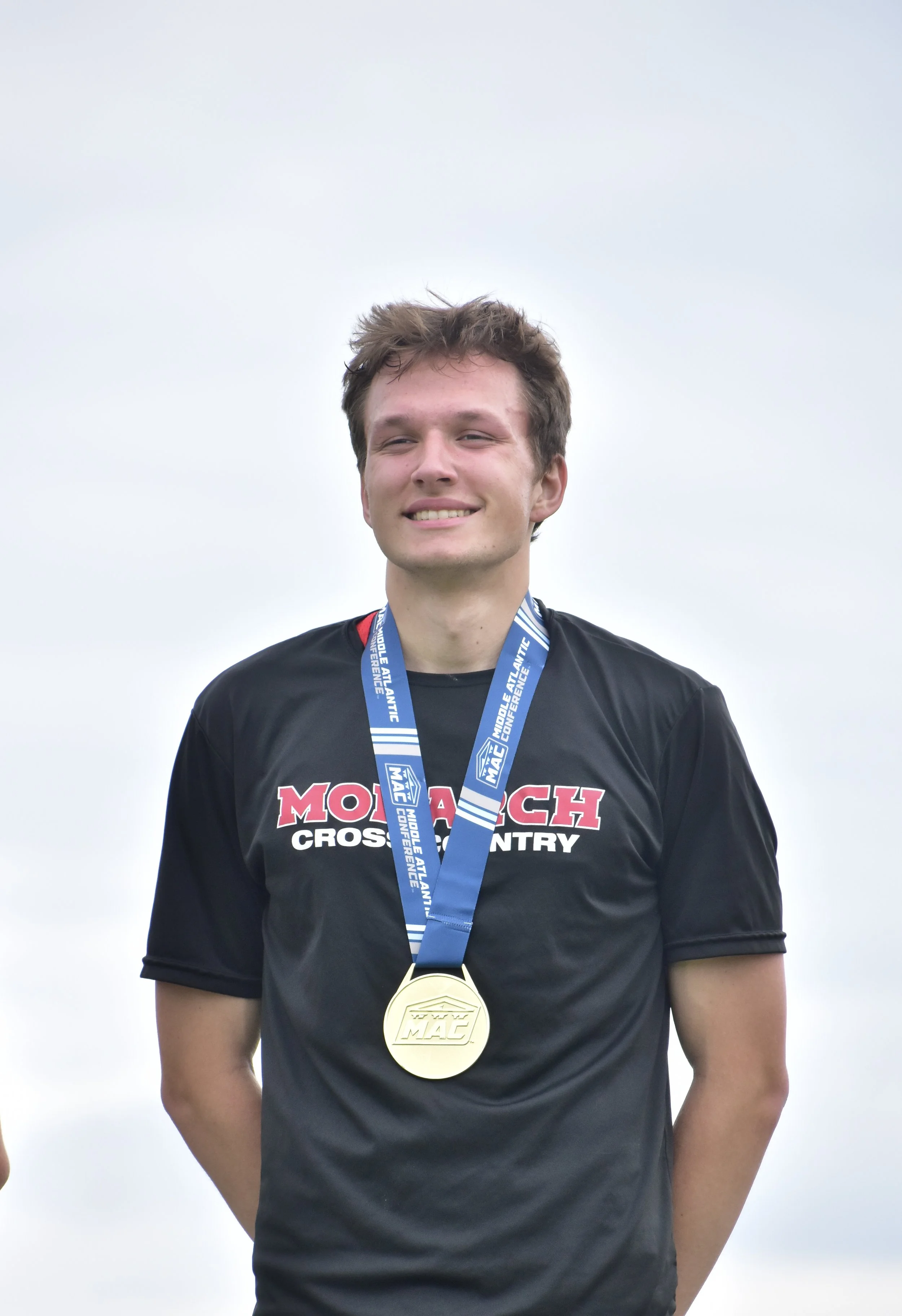 A young man with short brown hair smiling, wearing a black sports jersey with red and white text and a gold medal on a blue ribbon around his neck, standing outdoors under a cloudy sky.