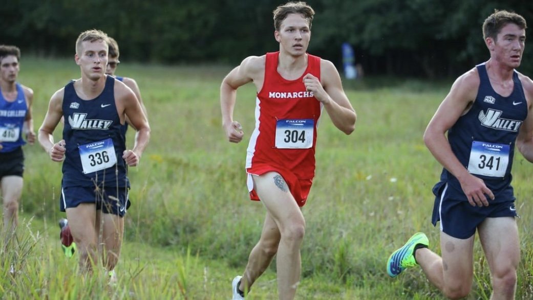 Male runners participating in a cross-country race on a grassy field, wearing athletic gear with race numbers.