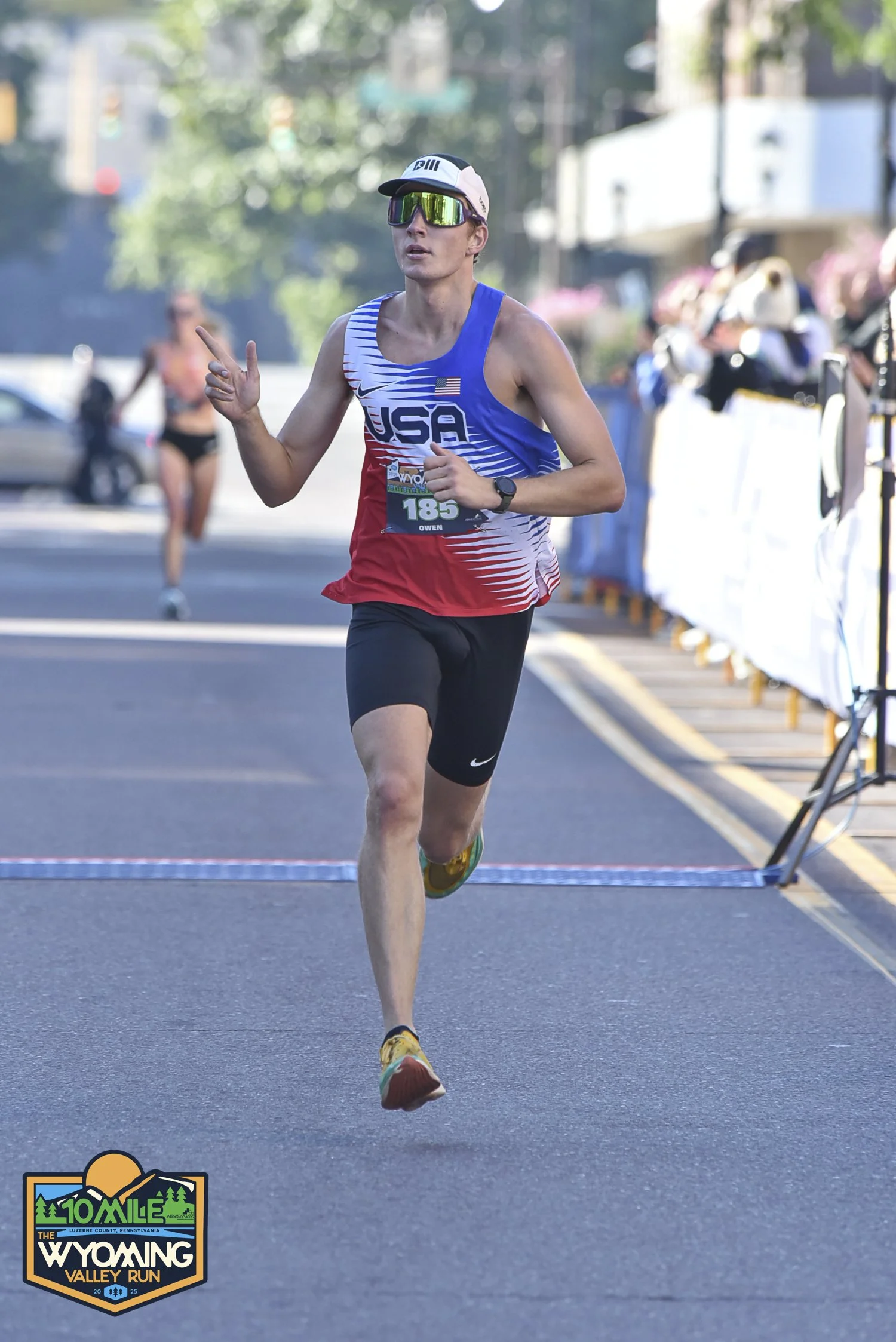 Male runner in USA race uniform crossing the finish line at the Wyoming Valley Run, Luzerne County, Pennsylvania, with other runners and spectators in the background.