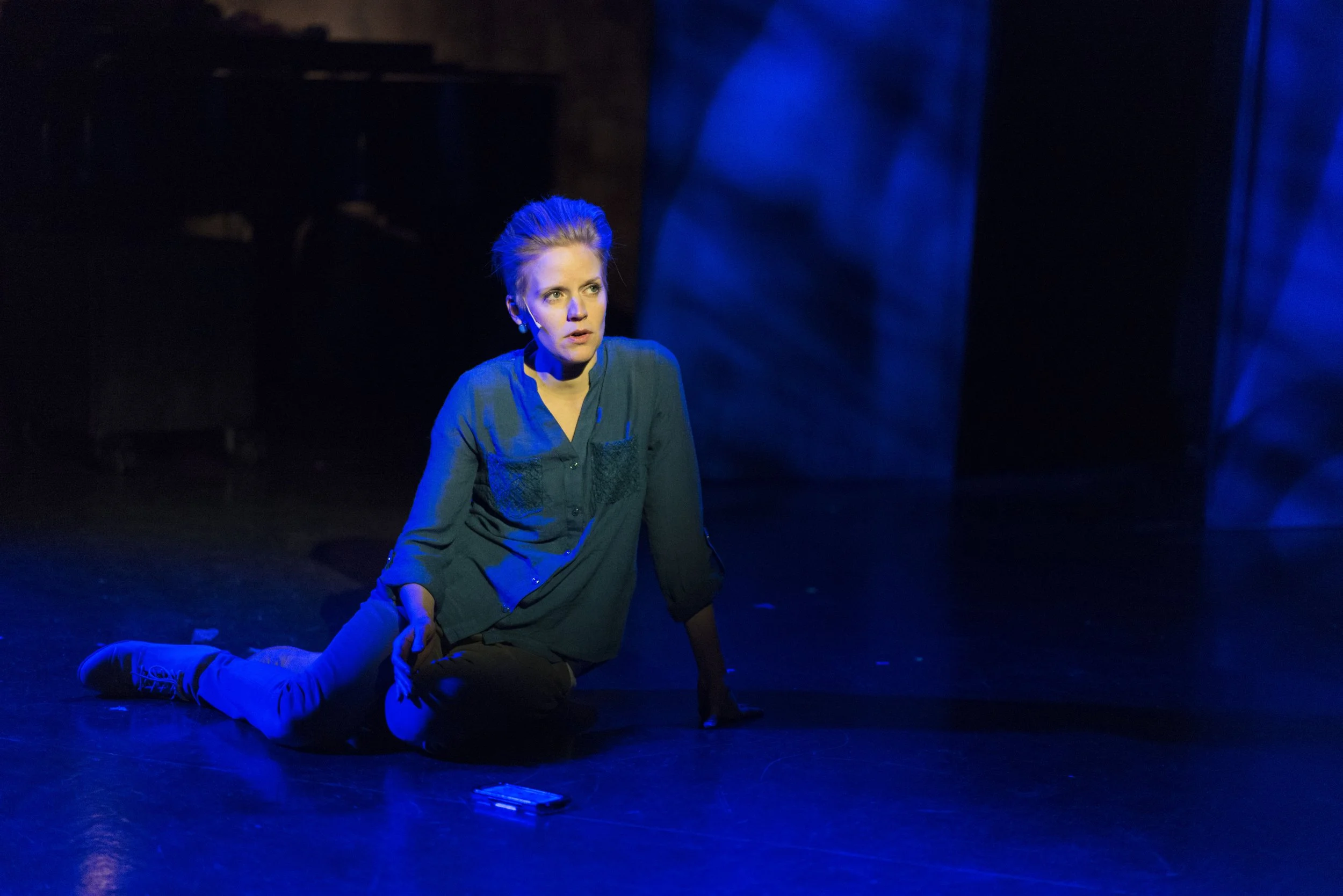 A woman with blonde hair styled up, wearing a dark shirt, is sitting on the floor in a dark room with blue stage lighting, looking up intently.