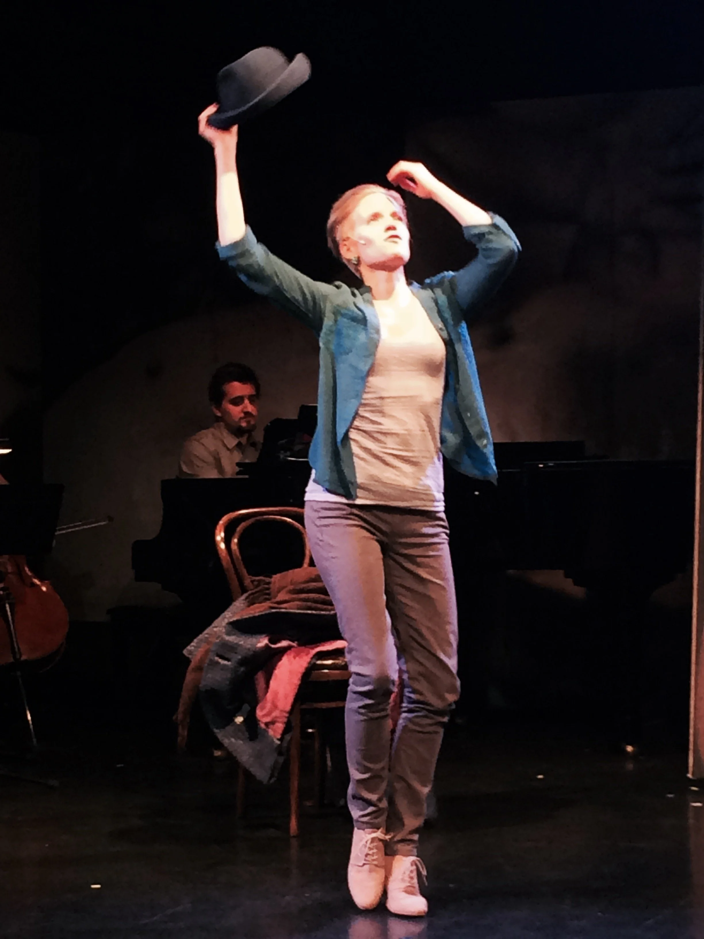 A woman on stage holding a hat with her right hand and looking upward. A man in the background is playing a piano. The stage is dimly lit with a dark background.