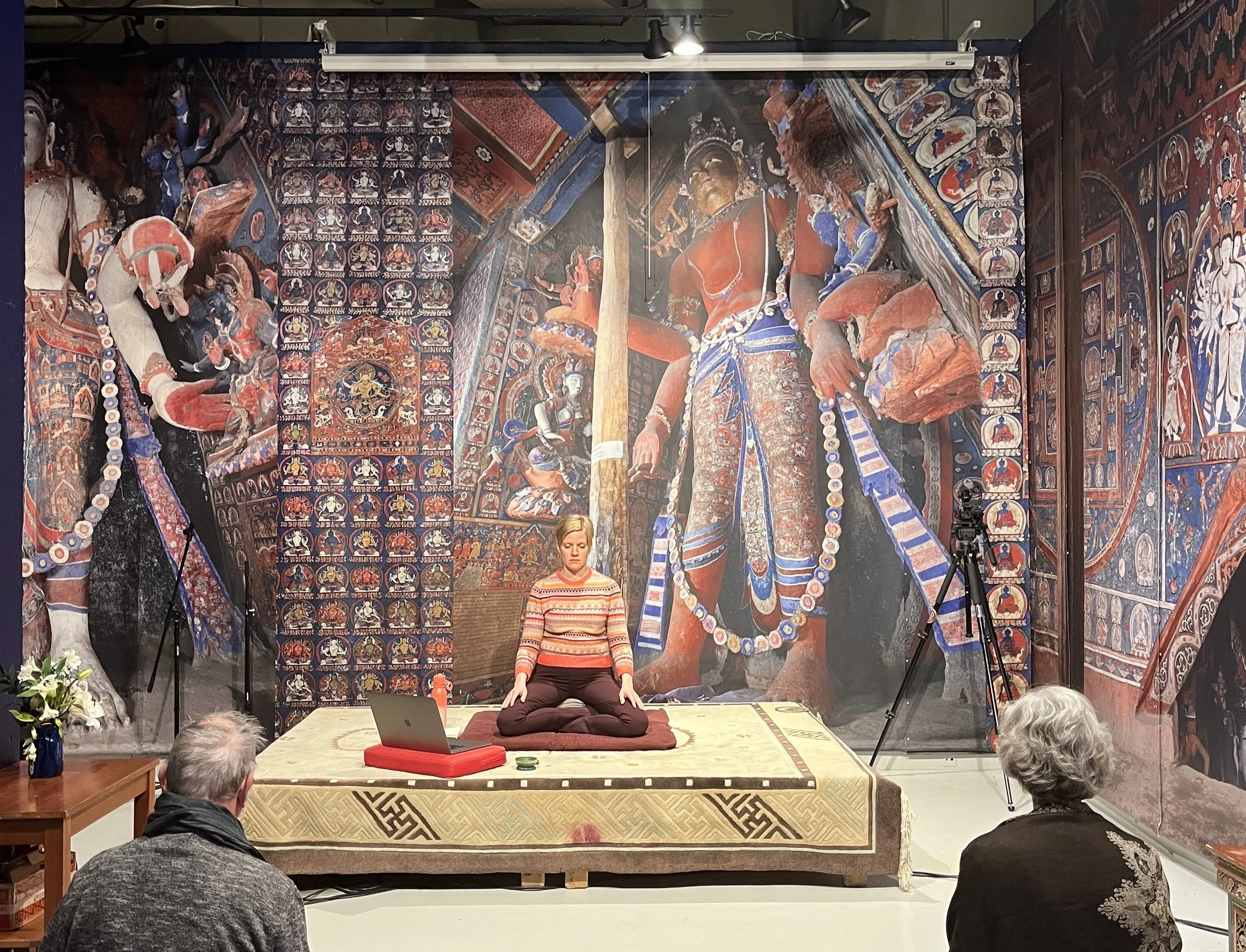 A woman practicing meditation on a stage in front of a large mural with traditional religious artwork. The woman is sitting cross-legged on a cushion, with her hands resting on her knees, eyes closed, and wearing a striped sweater. There is a laptop, a water bottle, and a microphone on the stage. Audience members are seated in front of the stage, watching. The mural behind features intricate depictions of deities and religious symbols in vibrant colors.