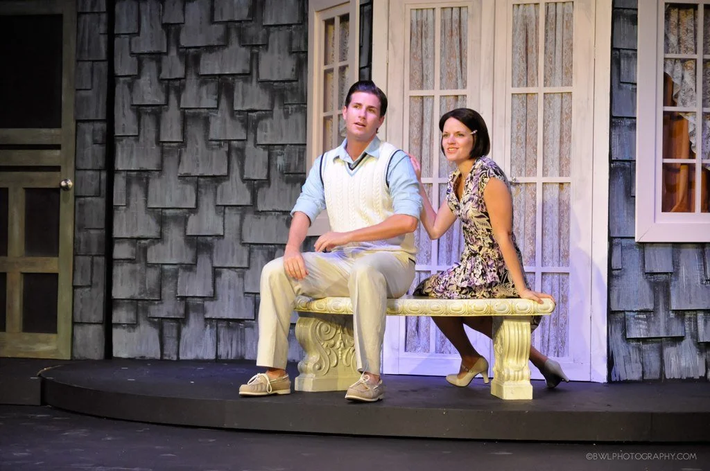 A man and a woman sit on a decorative bench on a stage, with the woman touching the man's shoulder.