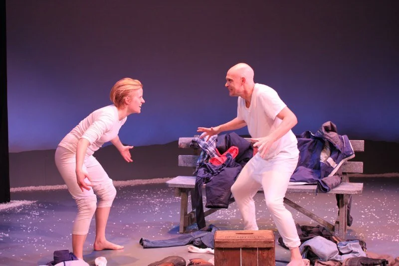 Two actors performing on stage, one woman and one man, with a bench and scattered clothes and props around them, in a theatrical setting.
